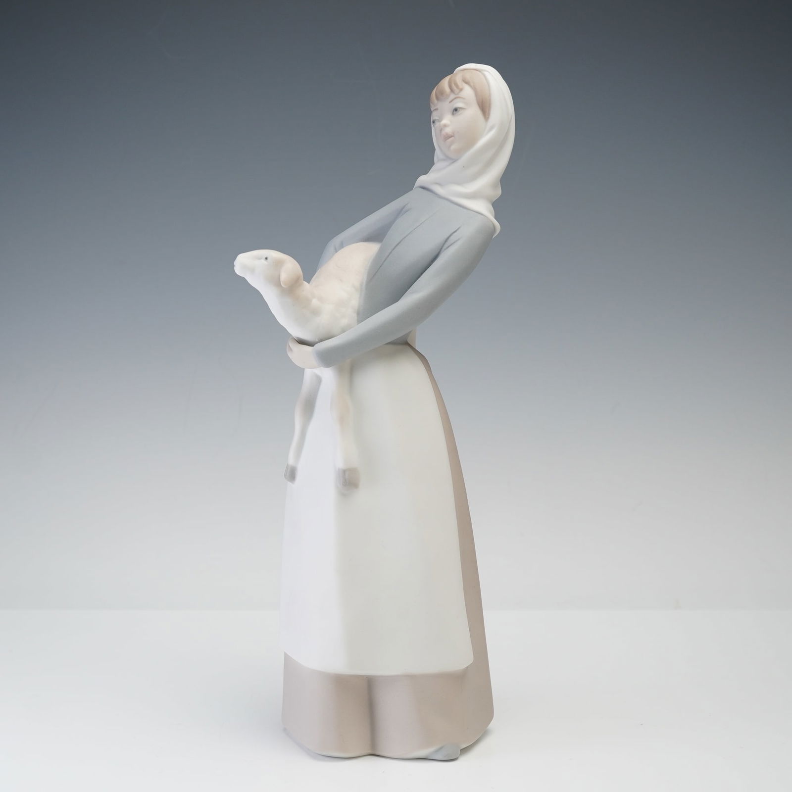 Lladro Porcelain Figurine, Girl with Lamb 1014584: A Lladro porcelain figurine titled Girl with Lamb, model number 1014584. The piece depicts a serene young woman in a pale blue blouse, long white apron, and soft beige skirt, holding a lamb in her arm