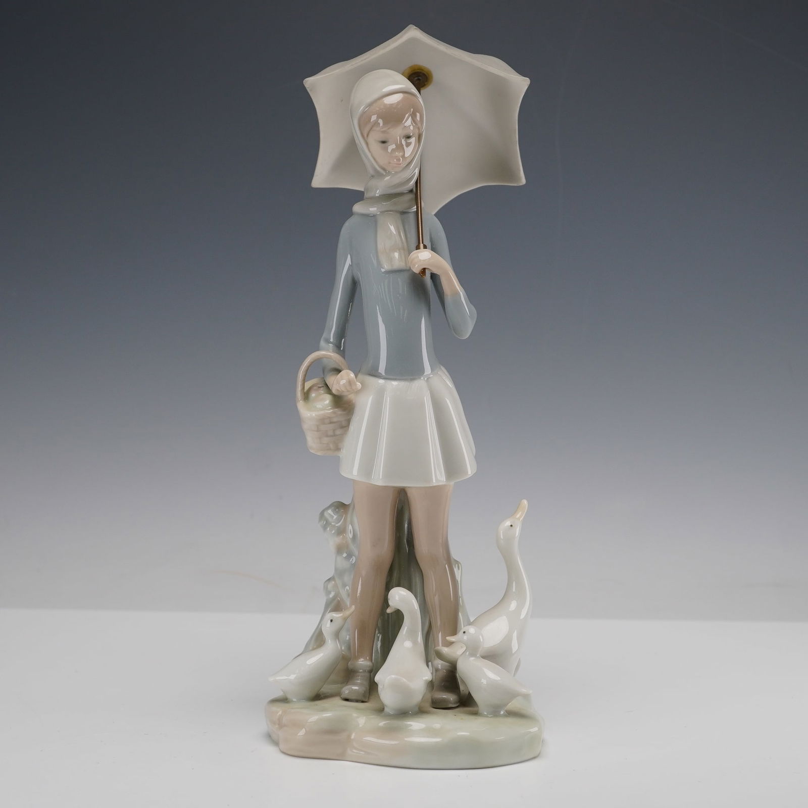 Lladro Porcelain Figurine, Girl with Umbrella and Geese 01004510: Figurine depicting a young woman holding an umbrella and carrying a basket as she stands surrounded by geese. Designed by Fulgencio Garcia, this delicate hand-painted sculpture features the brand's si