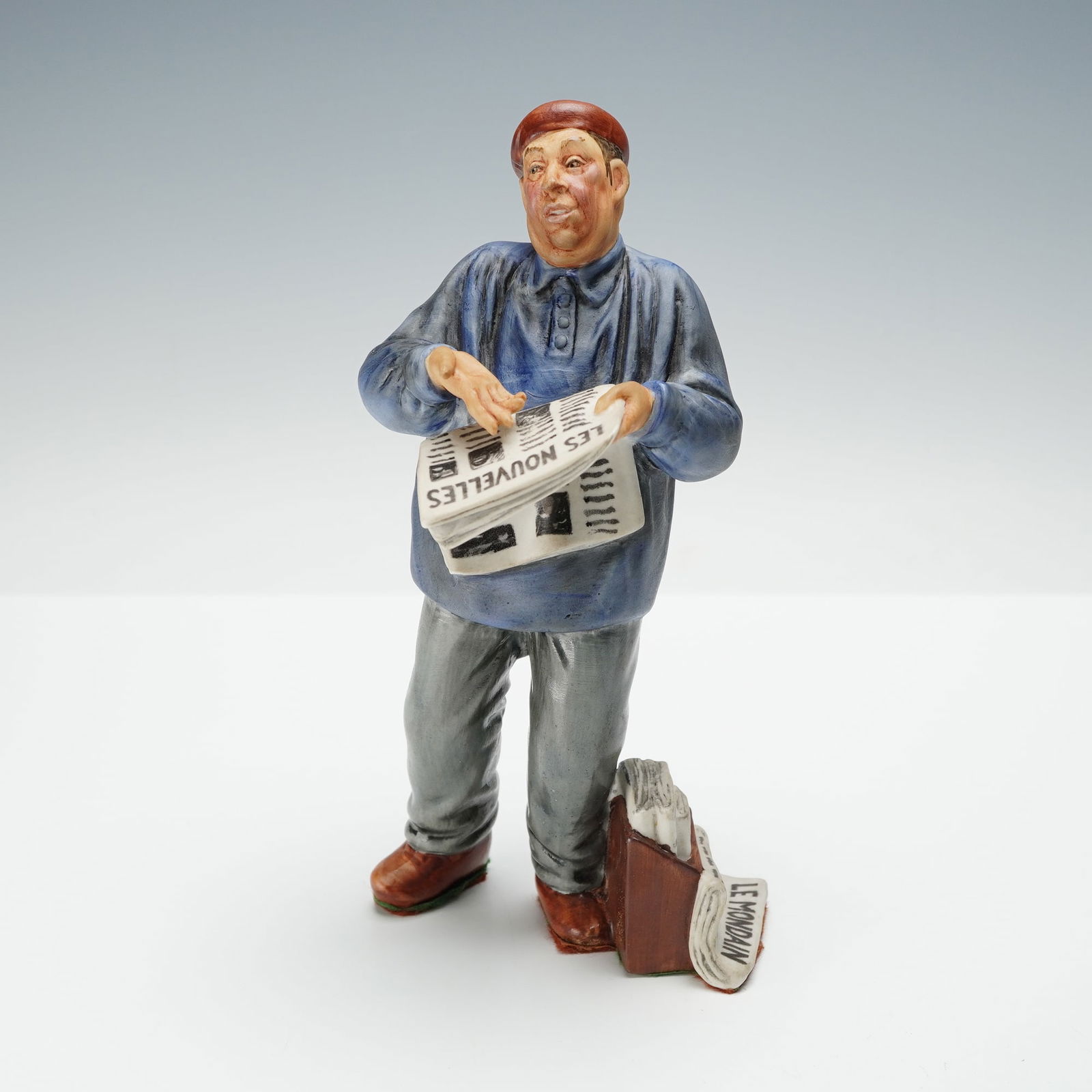 Royal Doulton Figurine, The Parisian HN2445: Hand-painted figurine from the Character Study was made in England and depicts a newspaper vendor wearing a blue shirt, grey trousers, and a red cap while holding a newspaper. A brown satchel with add