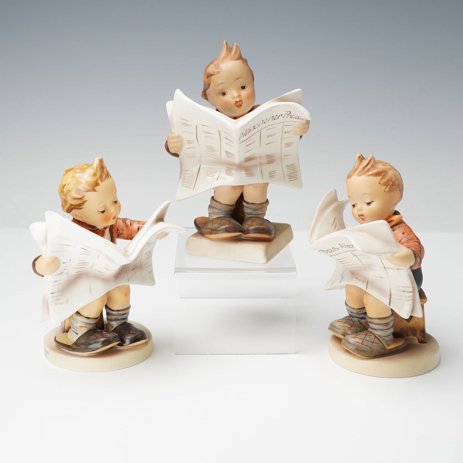 3pc Goebel Hummel Figurines, Latest News: Group of three Goebel Hummel porcelain figurines, each depicting a young boy reading a newspaper. The newspapers bear various German titles including Rüdesheimer Nachrichten, Wiesbadener Presse, and