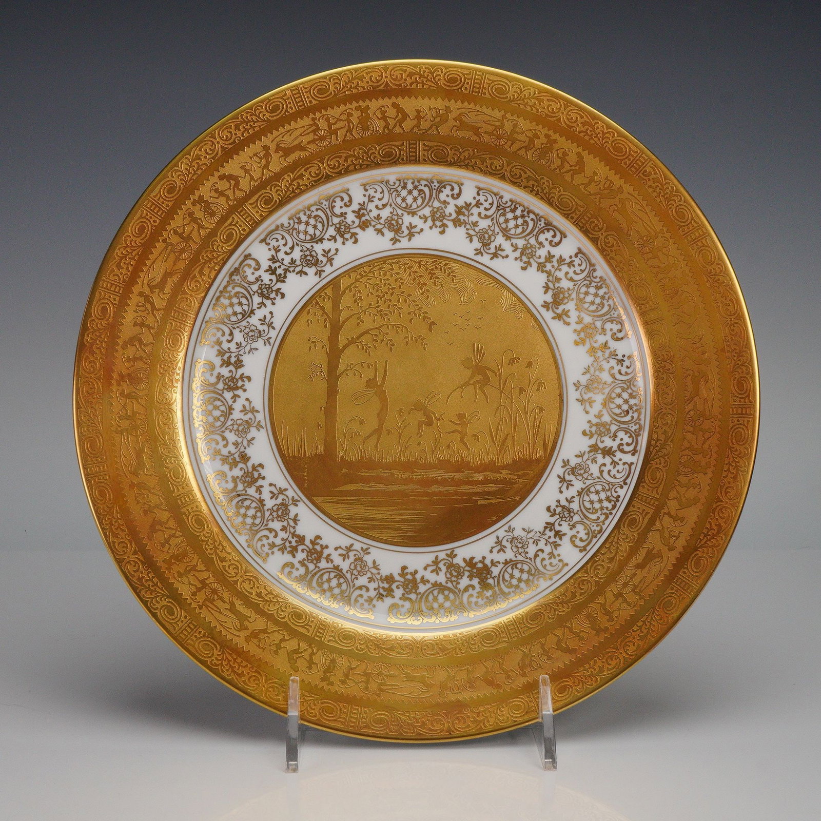 Gloria Fine Porcelain Acid-Etched Gold Dancing Fairies Decorative Plate: Glazed porcelain plate featuring an etched gilt central scene of dancing winged figures amid trees and foliage, surrounded by an ornate gilt foliate and figural border, marked on the underside with th