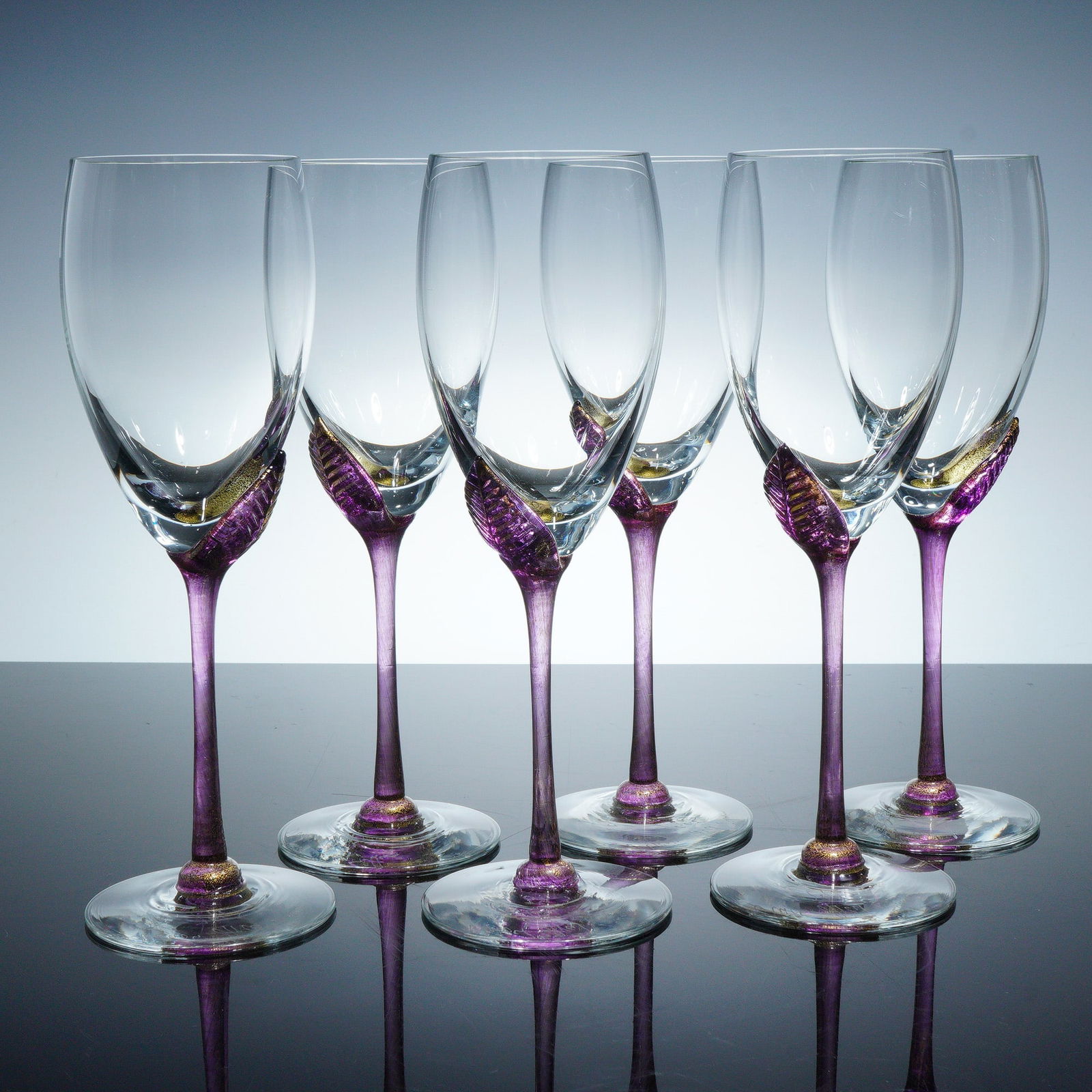 6pc Union Street Glass by Guy Corrie Wine Glasses: Six art glass goblets with clear bowls poised atop elongated stems of shimmering amethyst coloration, adorned with sculpted leaf motifs that wrap gracefully around the bowl's base. Signed "Union Stree