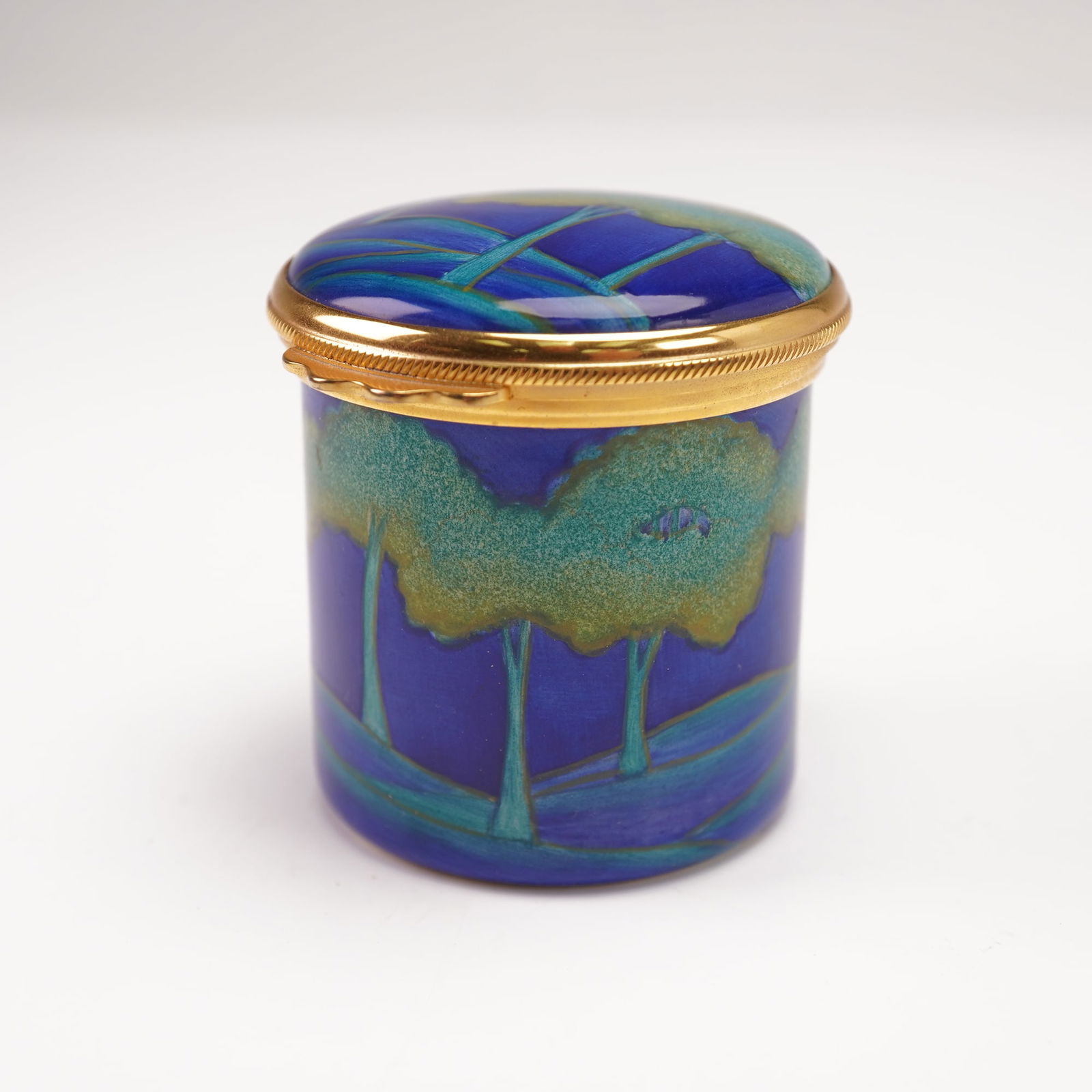 Moorcroft Enamels Cylindrical Pot, Moonlit Blue: Cylindrical lidded enamel pot in the "Moonlit Blue" design. The exterior decorated with stylized trees and rolling hills beneath a rich cobalt sky. It is fitted with gilt metal mounts and hinged cover