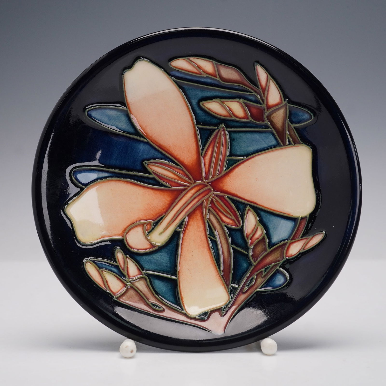 Moorcroft Pottery by Sian Leeper Coaster Dish, Panache: The circular dish features a stylized floral motif with raised tube lining and hand-painted glaze in shades of cream, orange, blue, and green on a dark ground. The underside is stamped Moorcroft with
