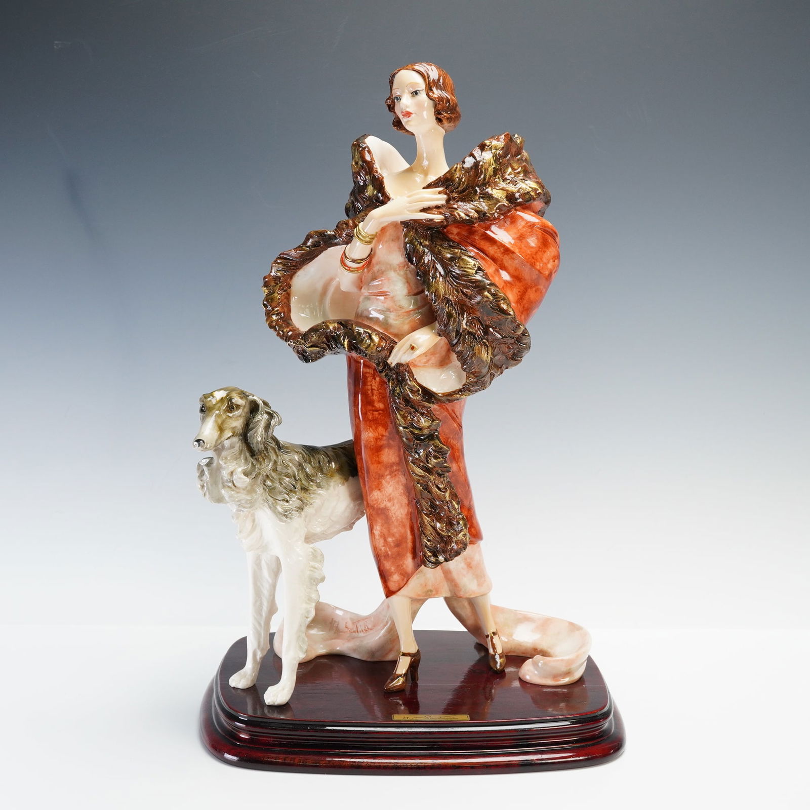 Capodimonte Amilcare Santini Sculpture, Woman with Borzoi Dog: A finely detailed porcelain sculpture depicting an elegant Art Deco-style woman in a flowing red fur coat, accompanied by a graceful Borzoi dog at her side. The figure stands on a polished wooden base