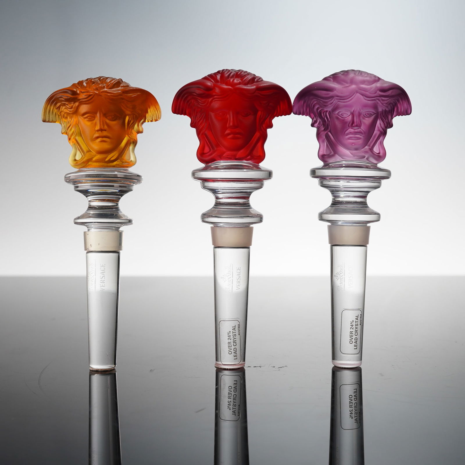 3pc Rosenthal Crystal Versace Medusa Bottle Stoppers- Amber, Red & Purple: Bottle stoppers from the Versace Medusa collection, showcasing richly colored Medusa heads in amber, ruby red, and amethyst purple frosted crystal. Each features the etched Rosenthal and Versace signa