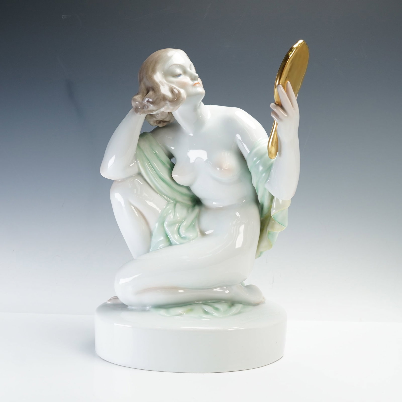 Herend by Szilagyi Nagy Istvan Porcelain Sculpture, Nude Woman with Mirror: A fine porcelain figure designed by artist Hungarian artist Szilagyi Nagy Istvan. The piece depicts a nude woman kneeling gracefully while gazing into a gilt mirror, partially draped in a soft green s