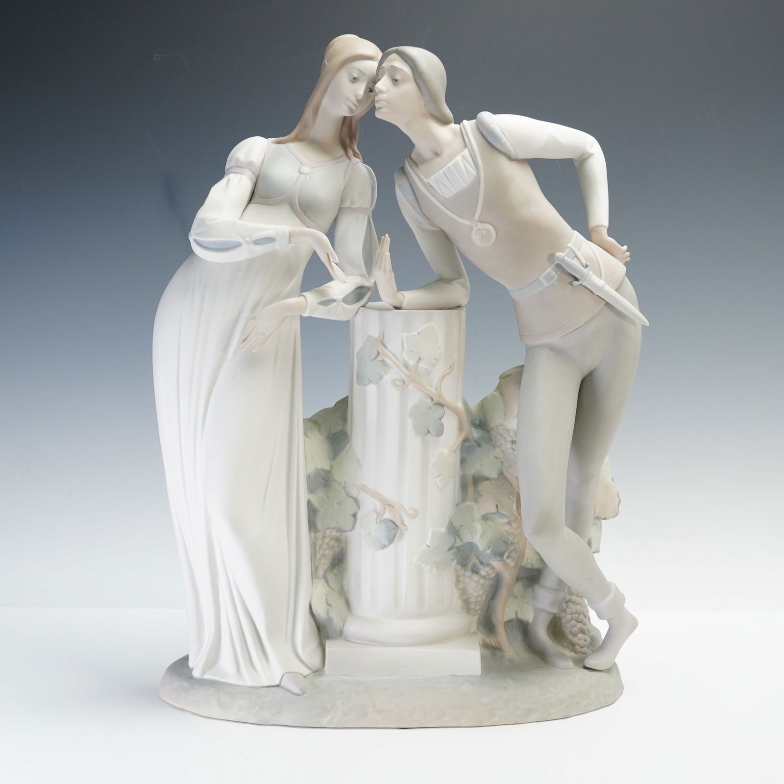 Lladro Porcelain Sculpture, Romeo and Juliet 1014750: A matte porcelain sculpture depicting the romantic scene of Romeo and Julie. The figures are modeled leaning toward one another over a classical column entwined with grapevines. Both characters displa