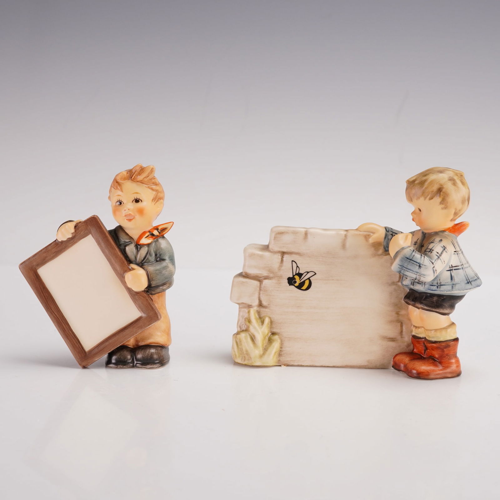 2pc Goebel Hummel Porcelain Figurines, Over the Horizon + Look What I Made: Charming Goebel Hummel porcelain figurines, both depicting young boys engaged in playful curiosity. Includes Over the Horizon (model 828) featuring a boy peering over a brick wall at a bee, and Look W
