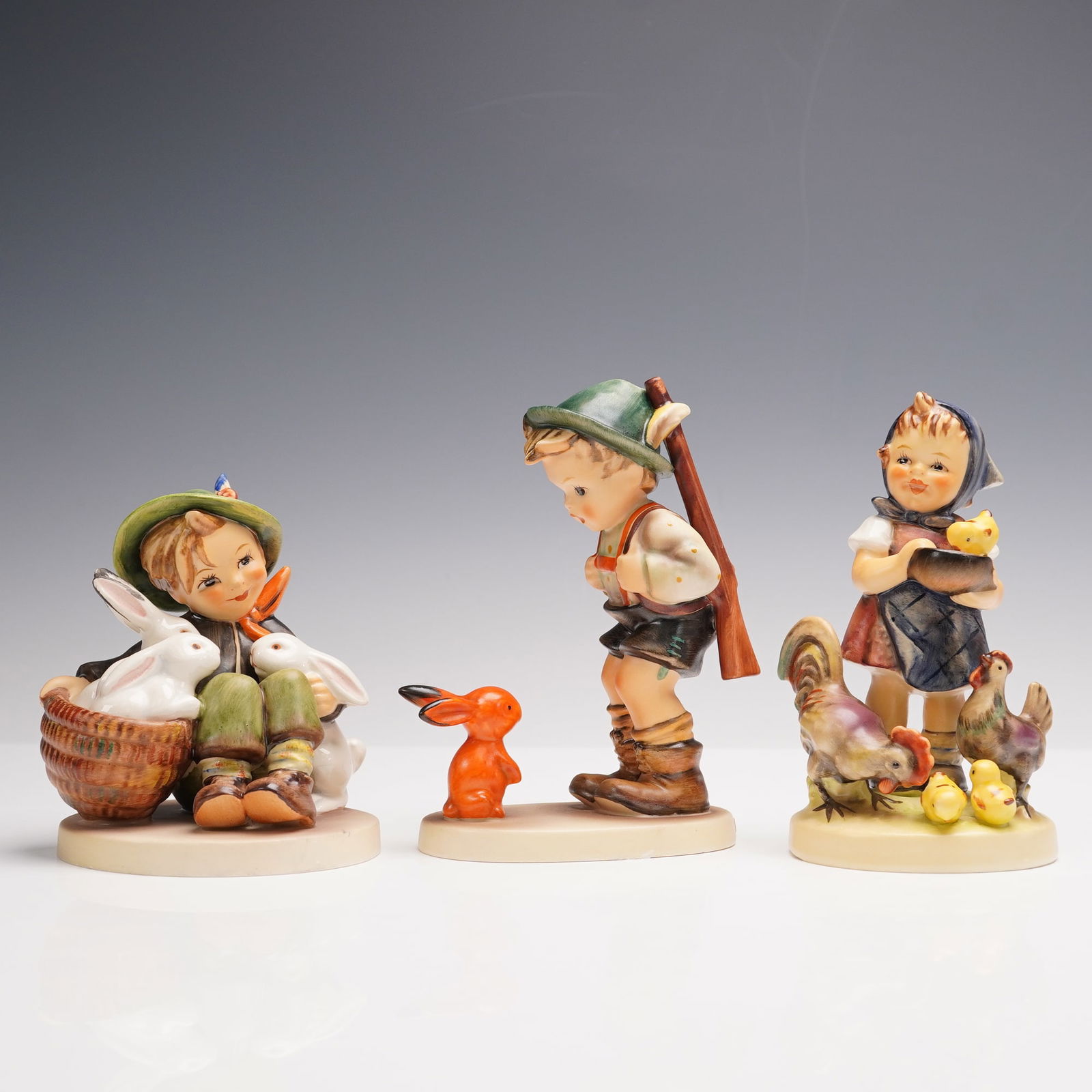 3pc Goebel Hummel Porcelain Figurines: A trio of Hummel porcelain figurines, each finely modeled and decorated with vibrant hand-painted detail. The grouping features Sensitive Hunter, Playmates, and Feeding Time. Various depictions such a