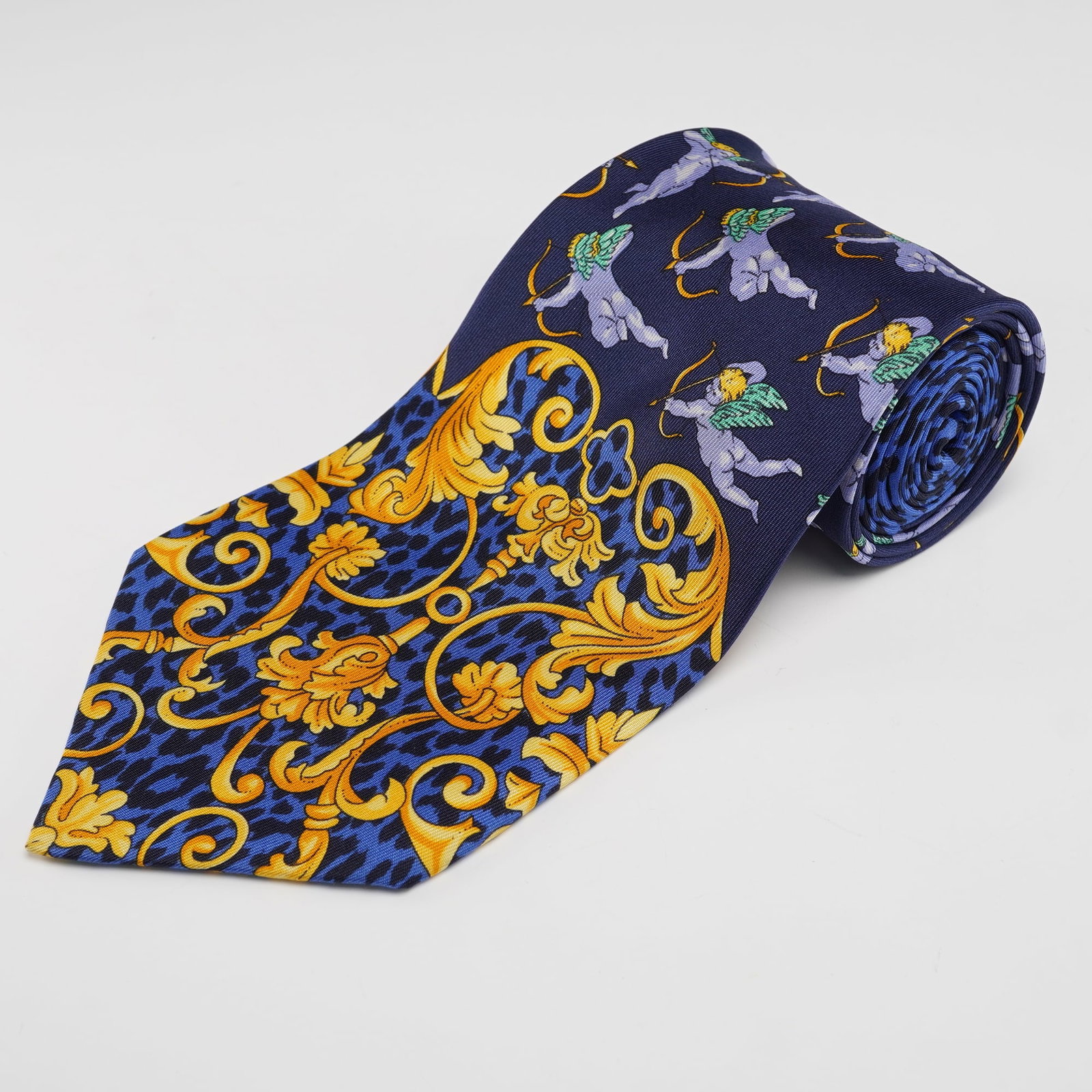 Gianni Versace Necktie, Dark Blue with Cherubs: This necktie is crafted in silk and features a detailed pattern that incorporates baroque-style scrolls in gold tones against a blue leopard-style ground. The design also includes small winged cherub