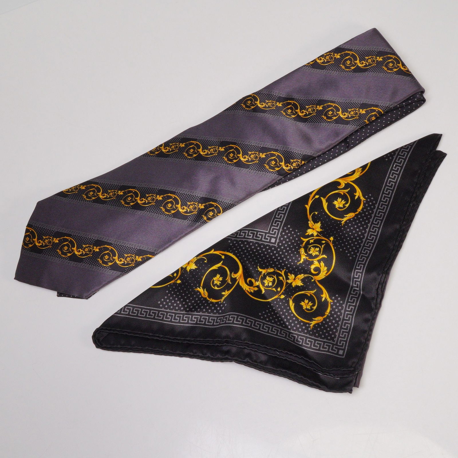 2pc Gianni Versace Necktie and Pocket Square Set: A Gianni Versace silk necktie and matching pocket square, both featuring the signature Atelier Versace Medusa motif and baroque scroll design in gold on black and gray backgrounds. The tie shows a dia
