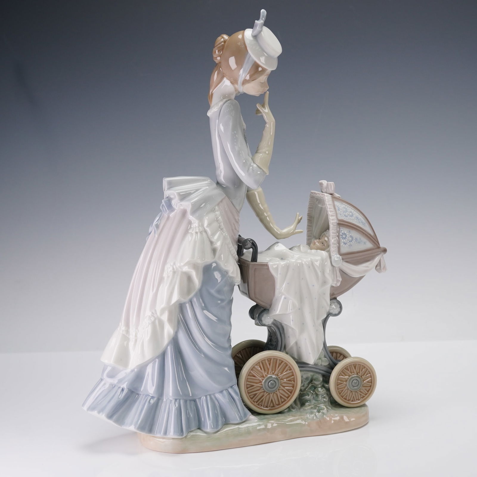 Lladro Porcelain Sculpture, Baby's Outing 01004938: Baby's Outing depicts a Victorian-era mother dressed in a ruffled pastel gown and small hat as she lovingly tends to her infant resting inside an ornate vintage pram. The stroller features a domed can