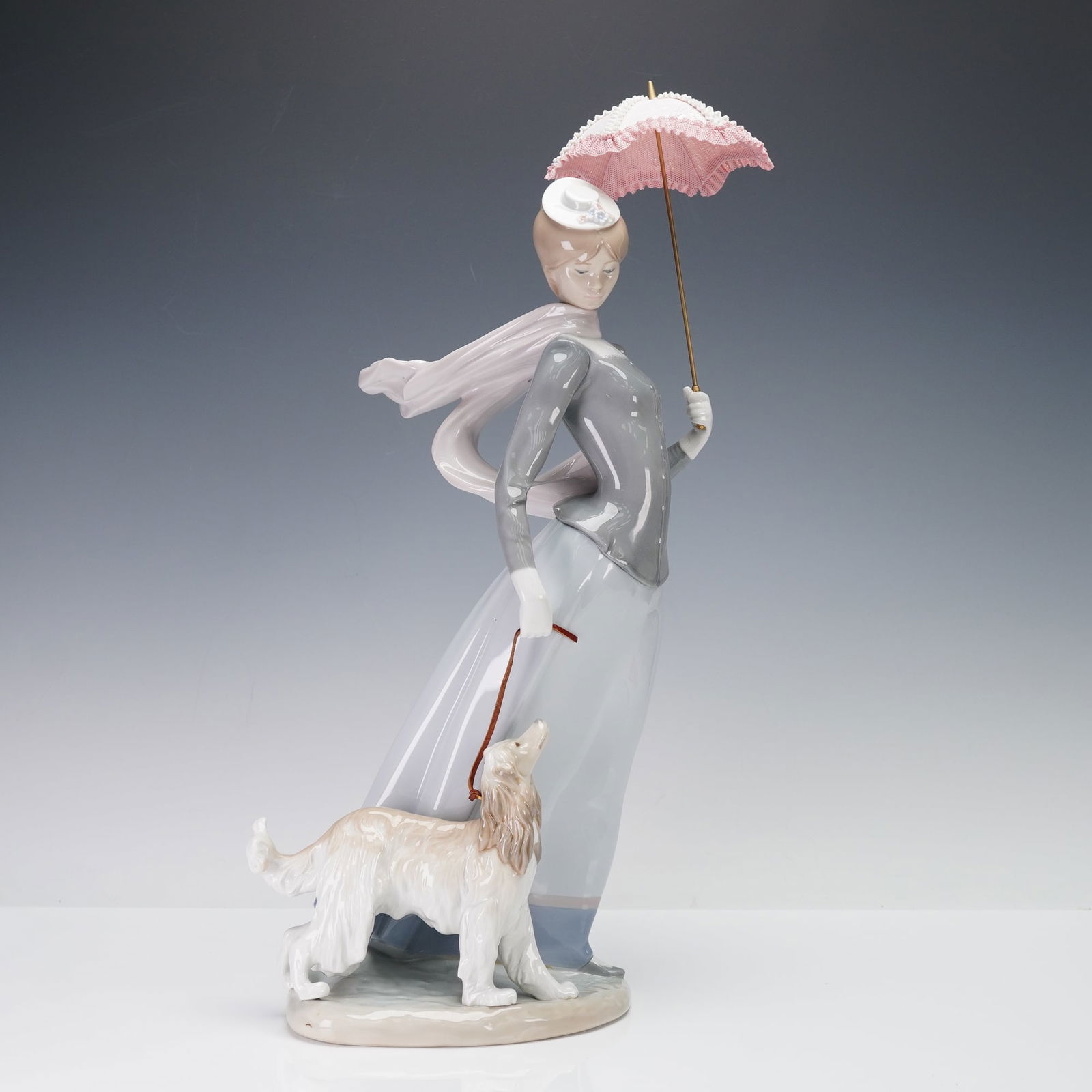 Lladro Porcelain Sculpture, Lady with Shawl 01008679: A woman in a flowing skirt and shawl holding a parasol while walking a long-haired dog, rendered in soft pastel glazes with detailed lacework on the umbrella and floral accent on her hat; underside be