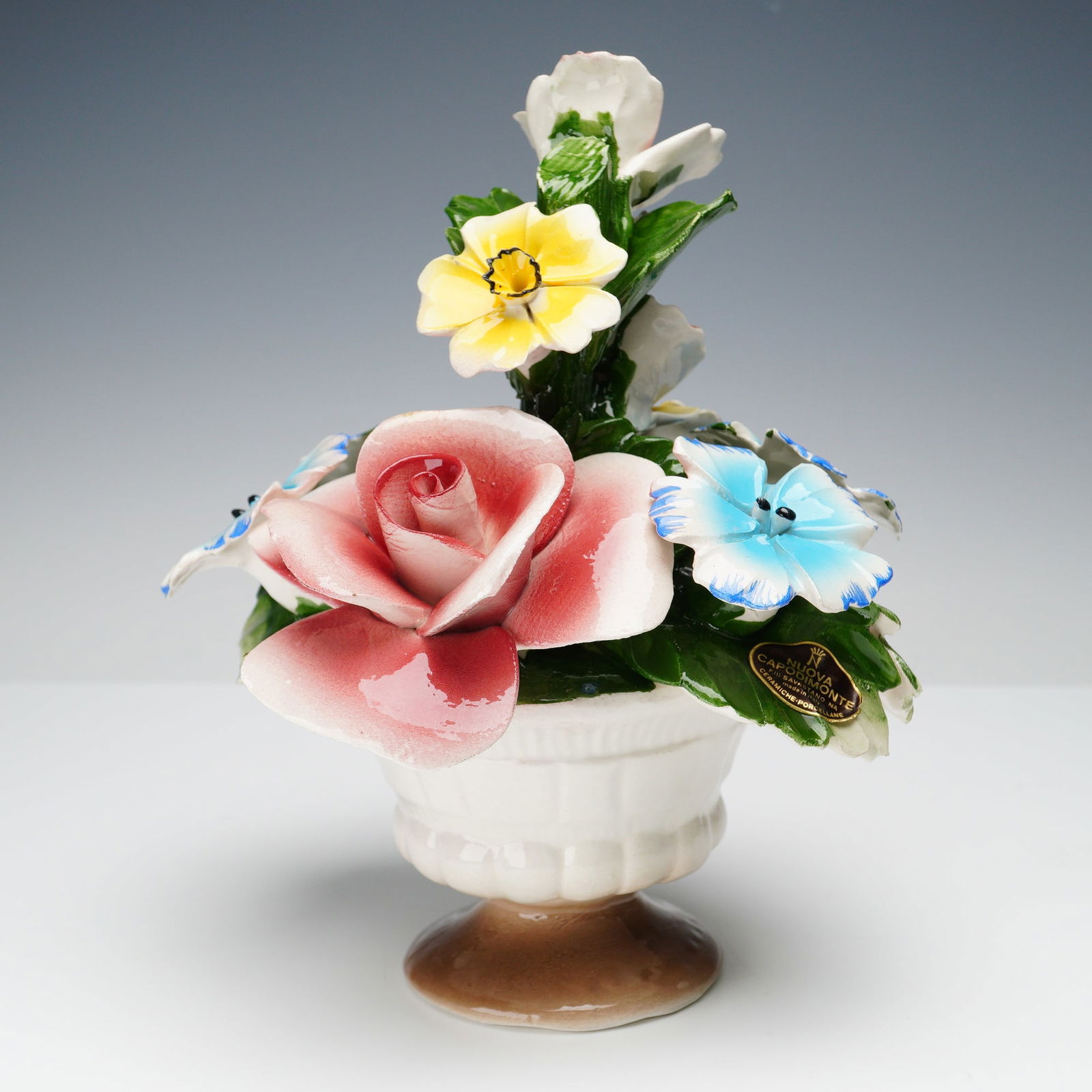 Nuova Capodimonte Porcelain Floral Arrangement Sculpture: Nuova Capodimonte porcelain floral bouquet in a pedestal vase, featuring a large pink rose surrounded by delicate blue, yellow, and white blossoms with green leaves. Beautifully hand-painted with high