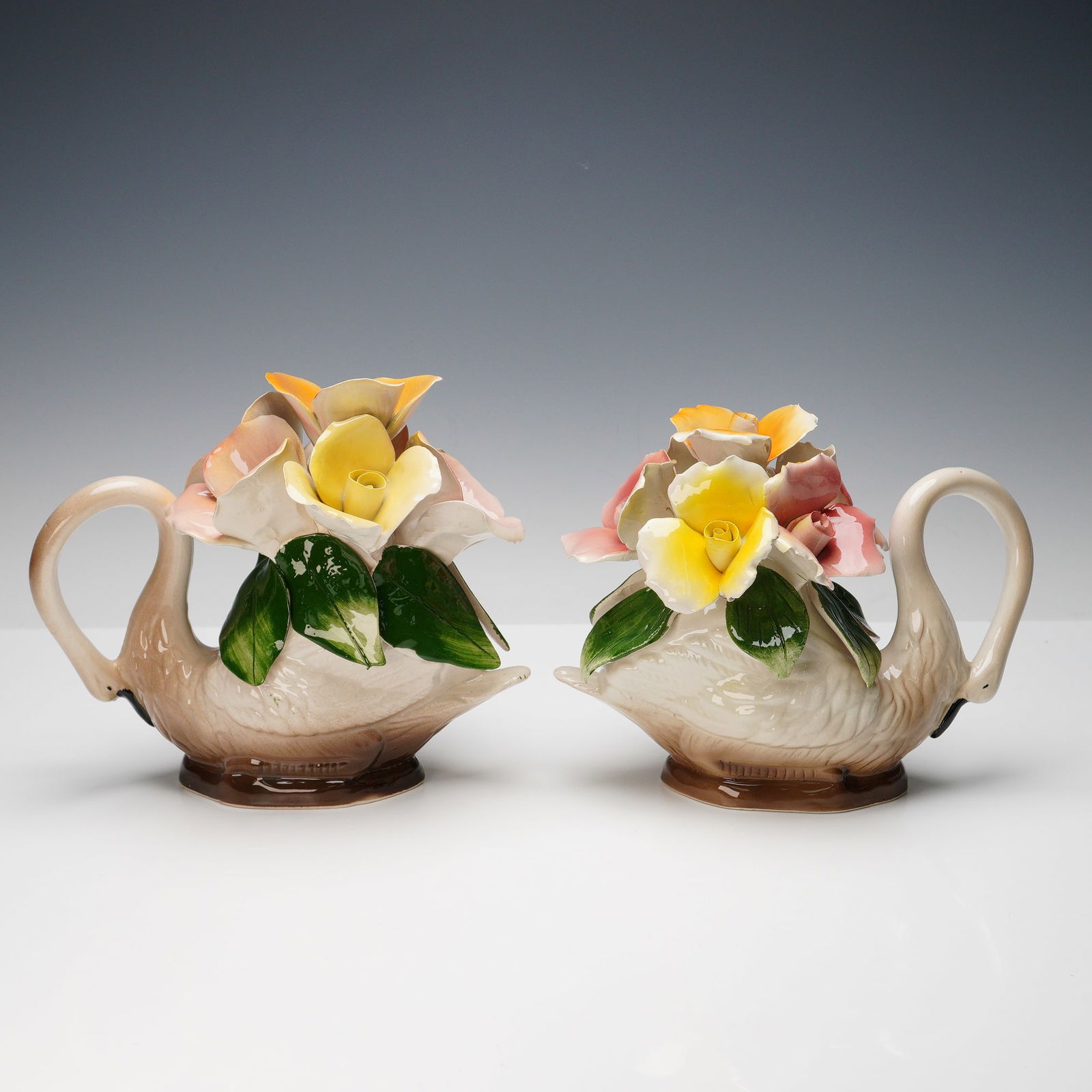 Pair of Nuova Capodimonte Decorative Floral Swan Figurines: Each beautifully crafted in ceramic with gracefully curved swan bodies supporting intricate hand-applied porcelain flowers in soft hues of yellow, pink, and peach with glossy green leaves. These elega