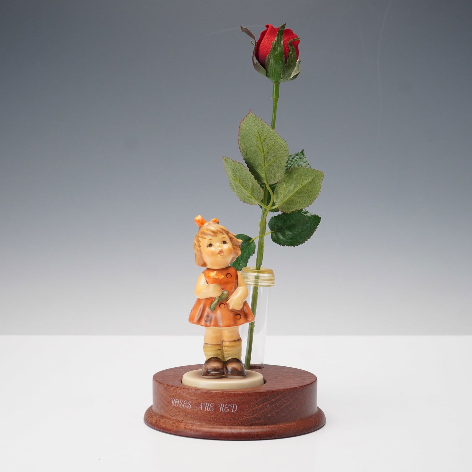 3pc Goebel Hummel Roses Are Red Figurine with Wooden Base and Rose: Delightful three-piece Goebel Hummel display set featuring a porcelain figurine of a young girl holding a flower, set on a wooden base engraved Roses Are Red. The base includes a removable glass bud v