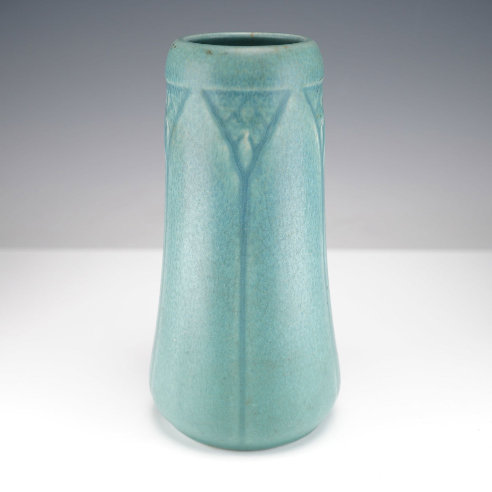Rookwood Pottery Turquoise Vase: This early twentieth century Rookwood Pottery vase features a tapered cylindrical form with subtle molded leaf and branch motifs beneath a soft matte blue glaze showing fine overall craquelure. The ba