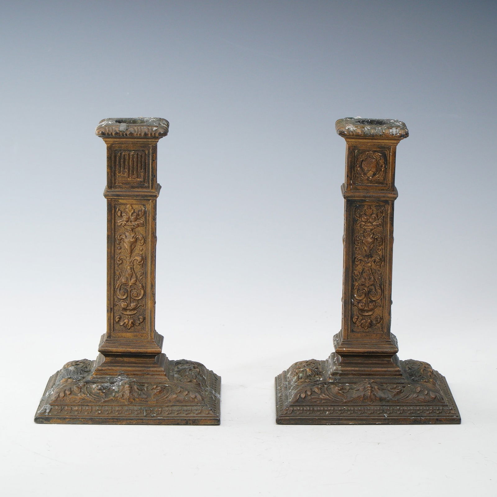 2pc Ornate Brass Candlestick Holders- Attributed to St. Louis VP Ball: A decorative pair of brass candlestick holders with square bases and columnar stems adorned with raised foliate and scroll ornamentation. The form and detailing resemble those attributed to the St. Lo