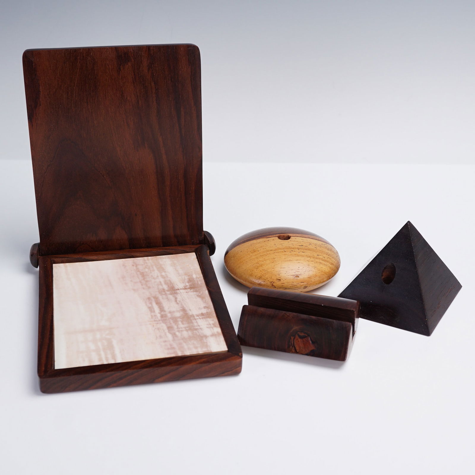 4pc Dark Wooden Desk Accessories, Pen Holders, Memo Pad, & More: Comprising a hinged memo pad holder with a light paper memo, a rounded two-tone wooden paperweight signed by the artist, a pyramid-shaped accessory with central opening, and a curved wooden pen rest.