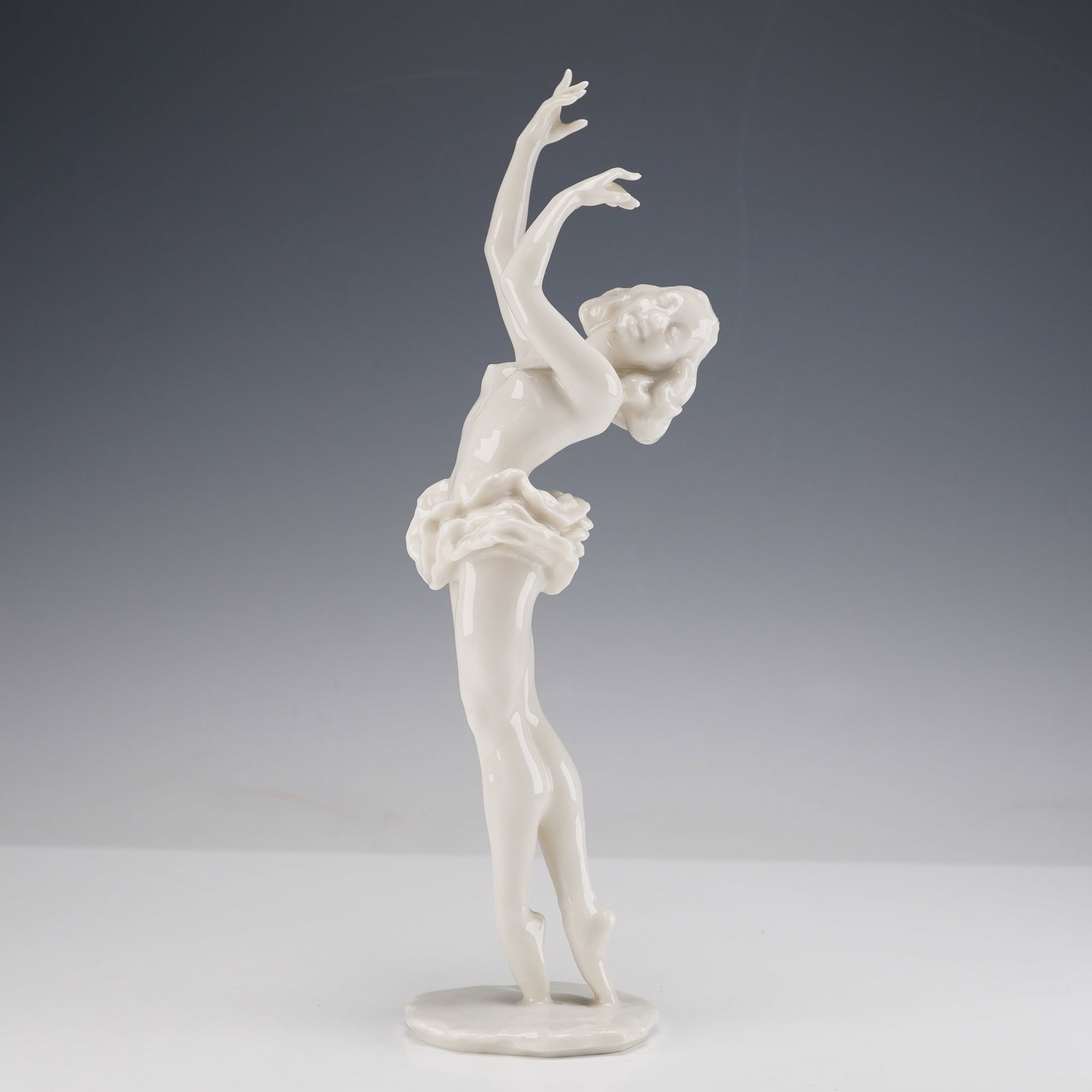 Hutschenreuther by Hans Achtziger Porcelain Ballerina Figurine: This white porcelain figurine depicts a ballerina balanced on one foot with her other leg extended behind her and both arms raised overhead. She is modeled in a short ruffled skirt and stands on a sha