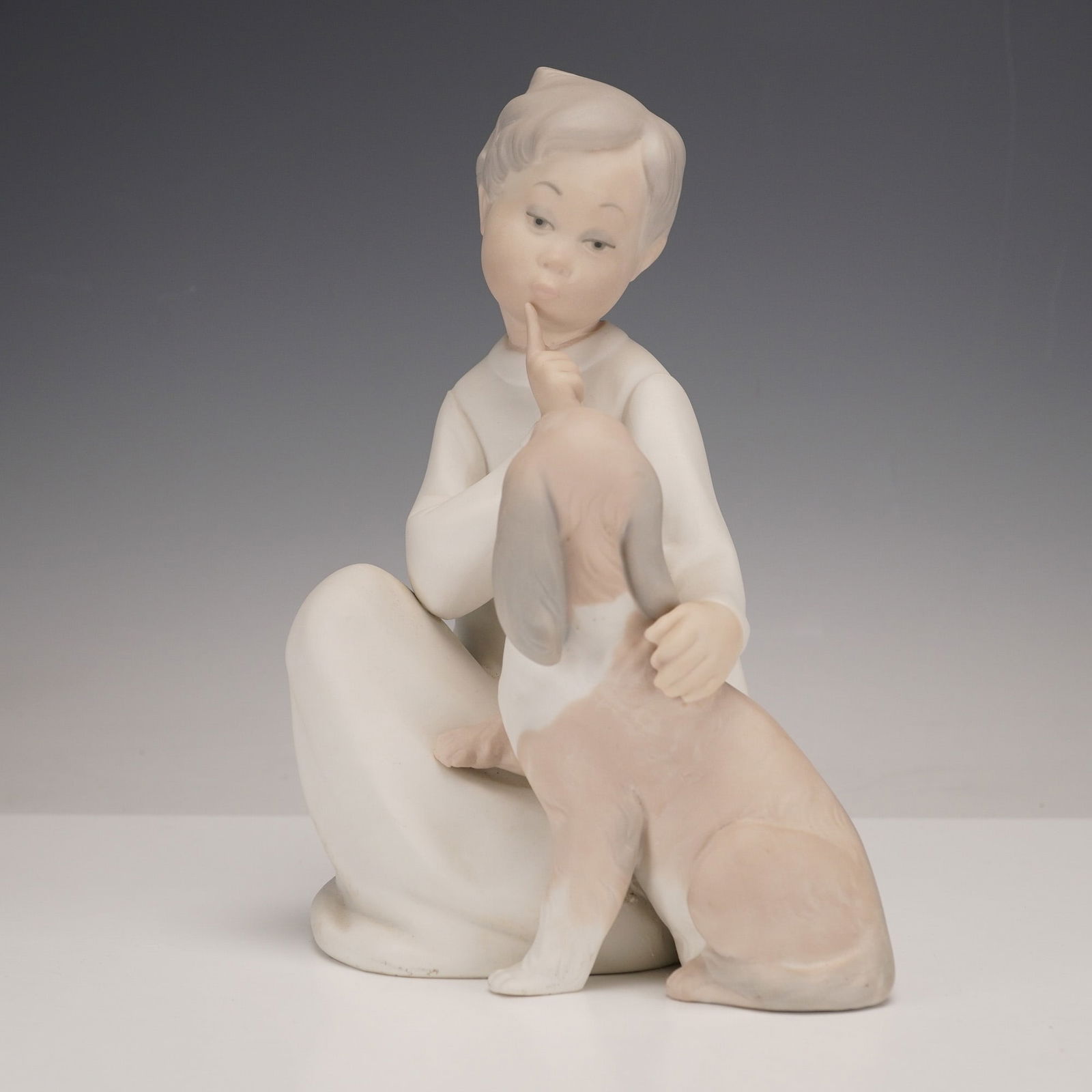 Lladro Matte Bisque Porcelain Figurine Boy with Dog #4522: A Lladro Boy with Dog figurine in matte bisque porcelain that depicts a seated child with one finger to their lips, affectionately interacting with a small dog looking up. The underside bears a blue s
