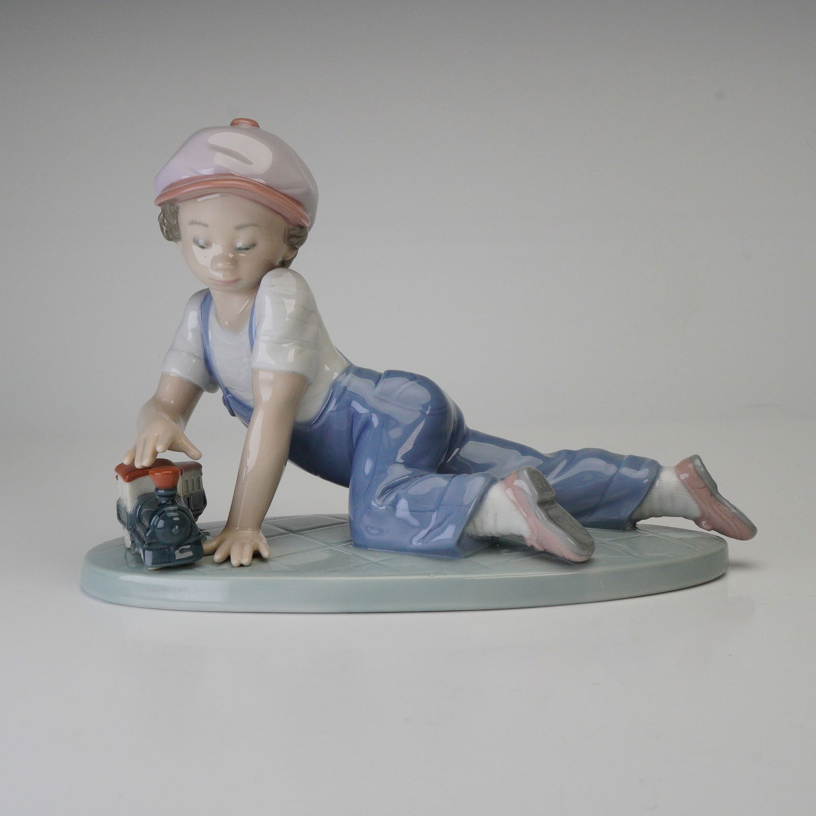 Lladro Porcelain Figurine, All Aboard 1007619: A 1993 Lladro Collectors Society issue. This glazed porcelain figurine depicts a young boy, dressed in a pink cap, white shirt, and blue overalls, kneeling and interacting with a small blue, red, and