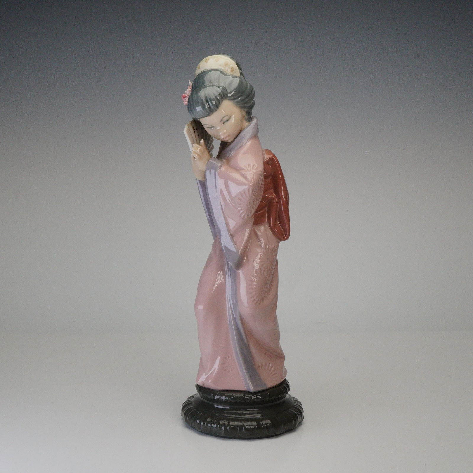 Lladro Porcelain Figurine, Chrysanthemum Geisha 1004990: A refined example of Lladro's celebrated artistry, Chrysanthemum Geisha 4990 portrays a young woman in a flowing pink and lavender kimono, delicately holding a decorative fan as she tilts her head in