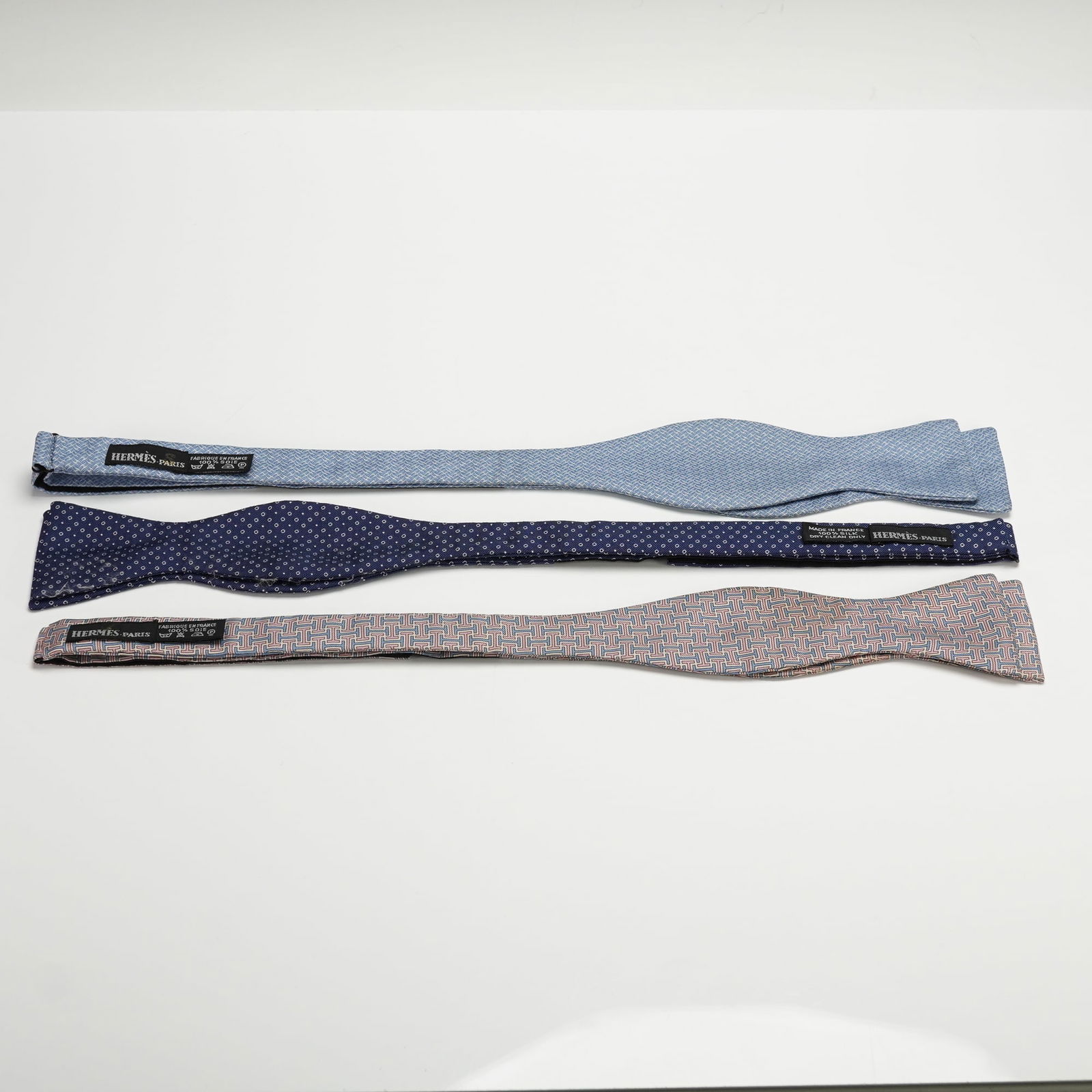 3pc Hermes Silk Bow Ties: This group includes three Hermes bow ties in assorted woven patterns. The set features a light blue geometric design, a navy dot pattern, and a multicolor geometric style. Each bow tie is made of 100
