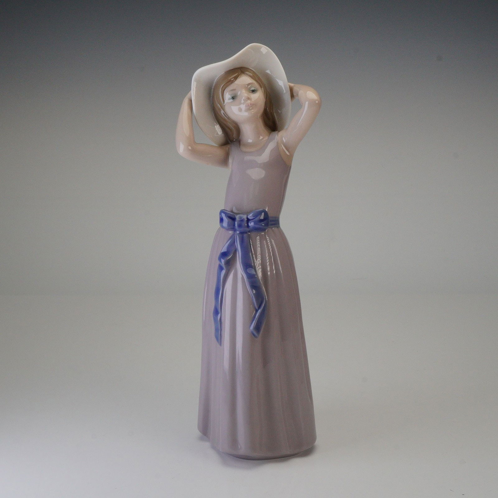 Lladro Figurine Coy Girl with Hat, Model 5011: A Lladro porcelain figurine, model number 5011, titled Coy and also known as Girl with Hat. This figurative piece depicts a young woman or girl holding a wide-brimmed white hat, dressed in a long, lig