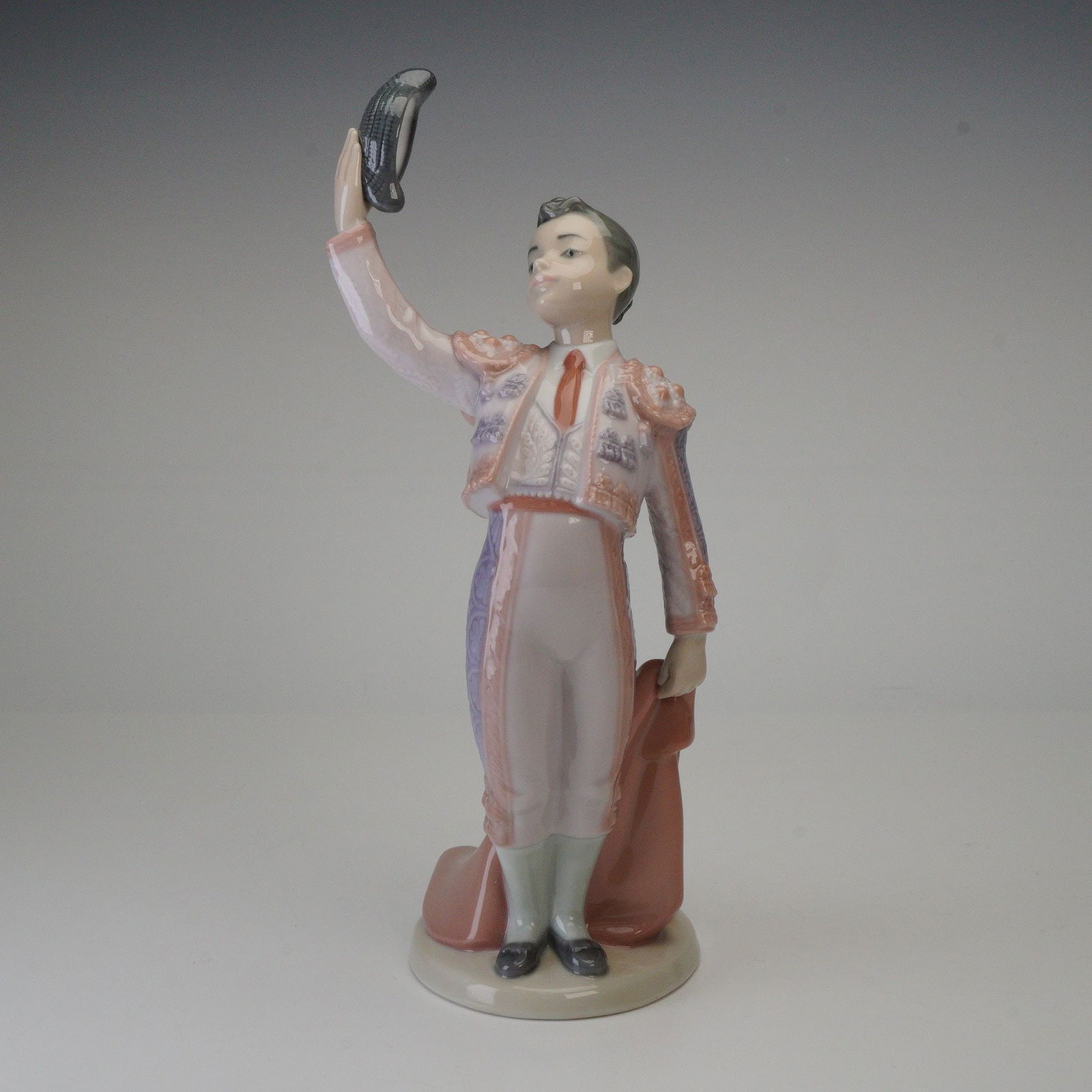 Lladro Porcelain Figurine, Little Matador 1006178: This glazed porcelain piece depicts a young male bullfighter in a traditional traje de luces uniform. The figure is posed holding a dark, textured montera hat aloft in his right hand, with a draped re