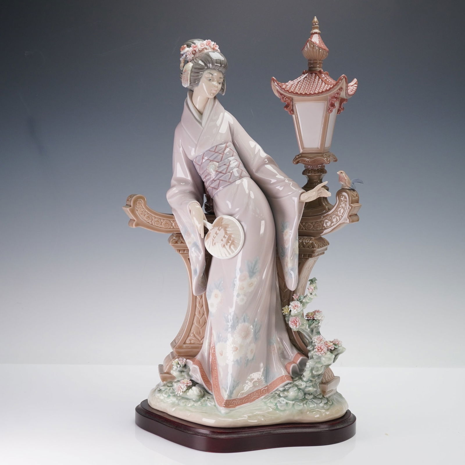 Lladro Porcelain Sculpture with Base, Mariko 01001421: A kimono-clad woman standing beside an ornate architectural balustrade and lantern, detailed with floral accents, a hand fan, and a small bird; includes the Lladro blue stamp and logo underside. This