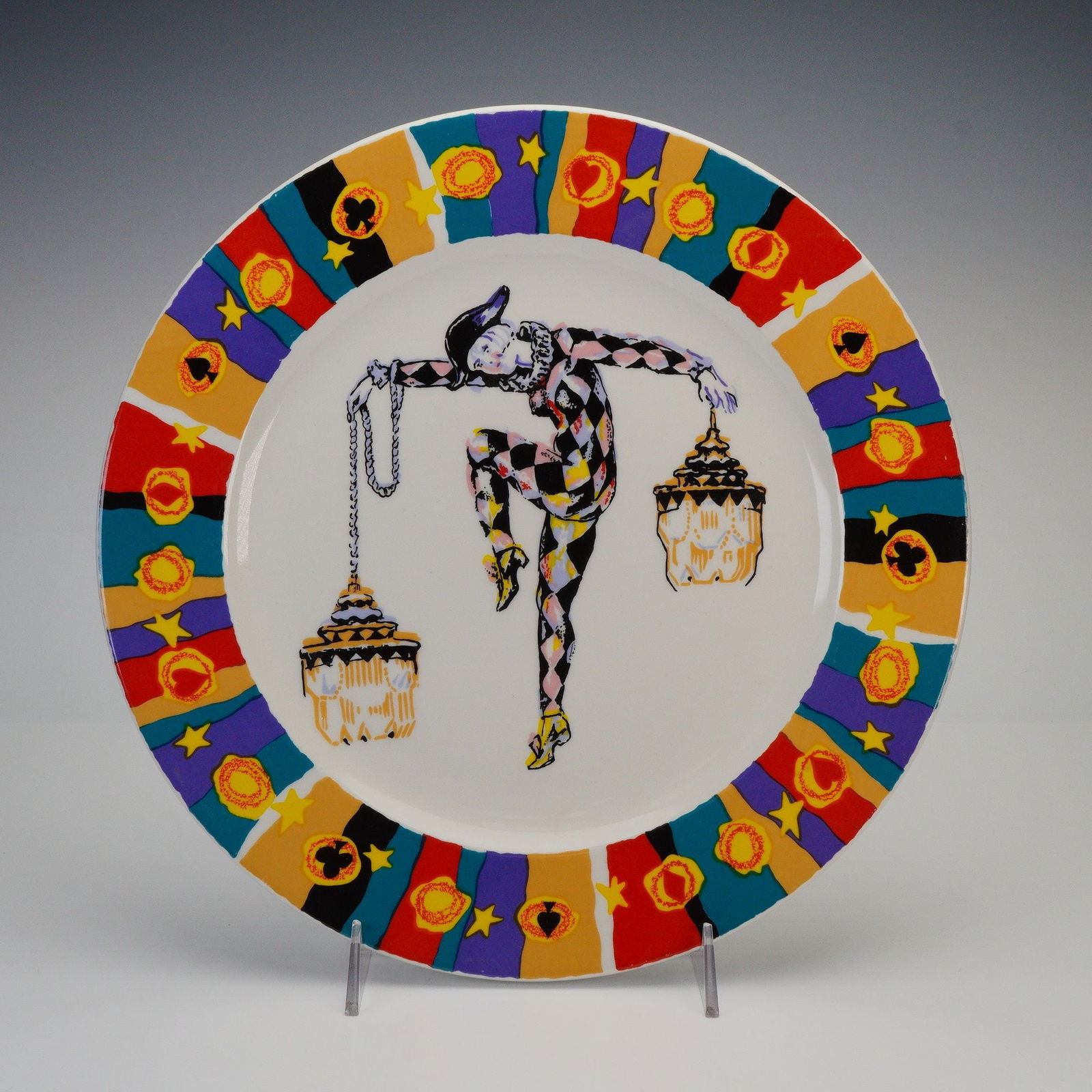Continental China Ceramic Harlequin Plate, Supra Dura: Decorative plate featuring a central depiction of a harlequin figure holding two hanging lanterns, surrounded by a wide rim decorated with colorful geometric panels, stars, and emblem-like motifs; und