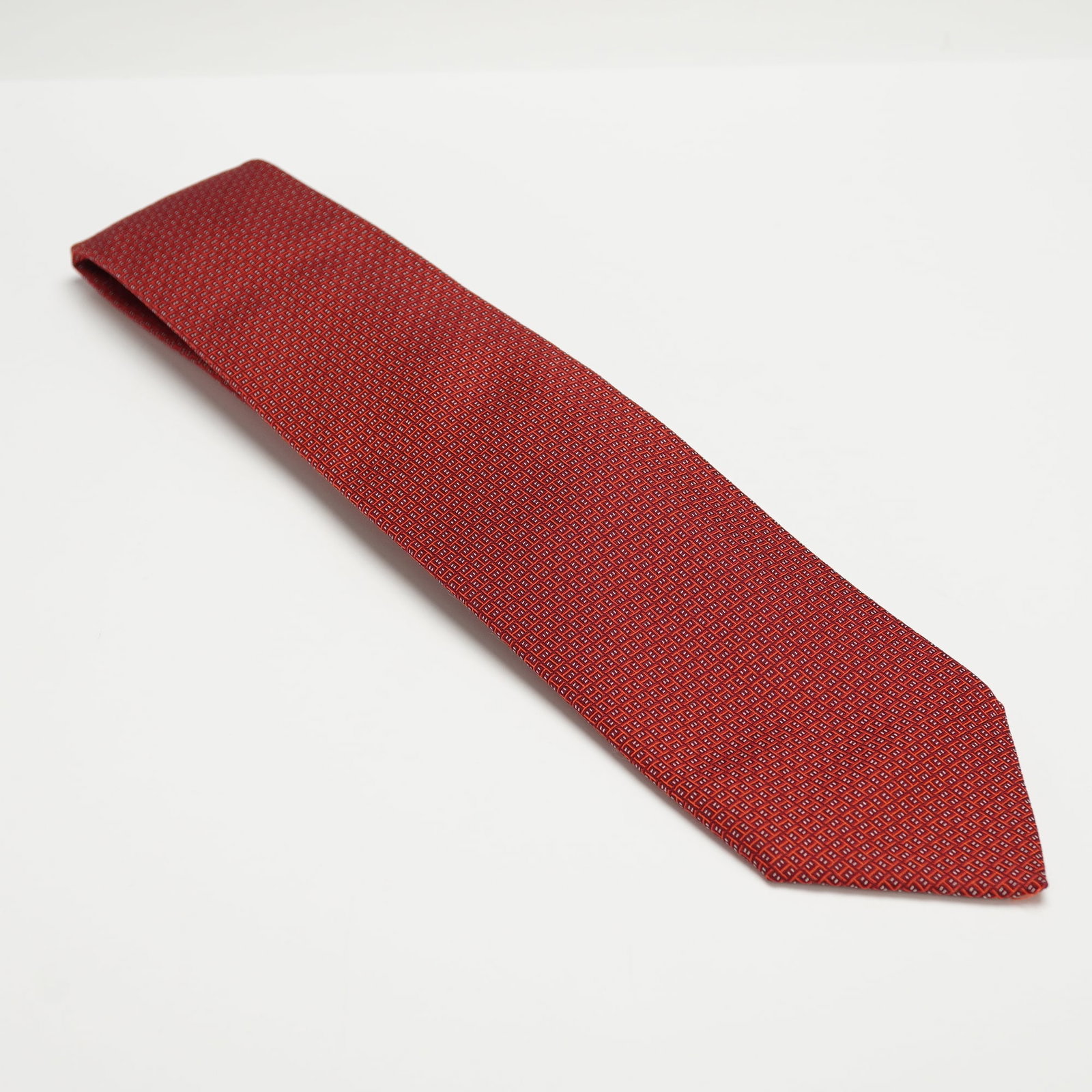 Hermes Red Geometric Pattern Necktie: This necktie features a repeating geometric pattern in red with small white accents. It is made of 100 percent silk and manufactured in France. The underside includes the Hermes-Paris label. Dimension