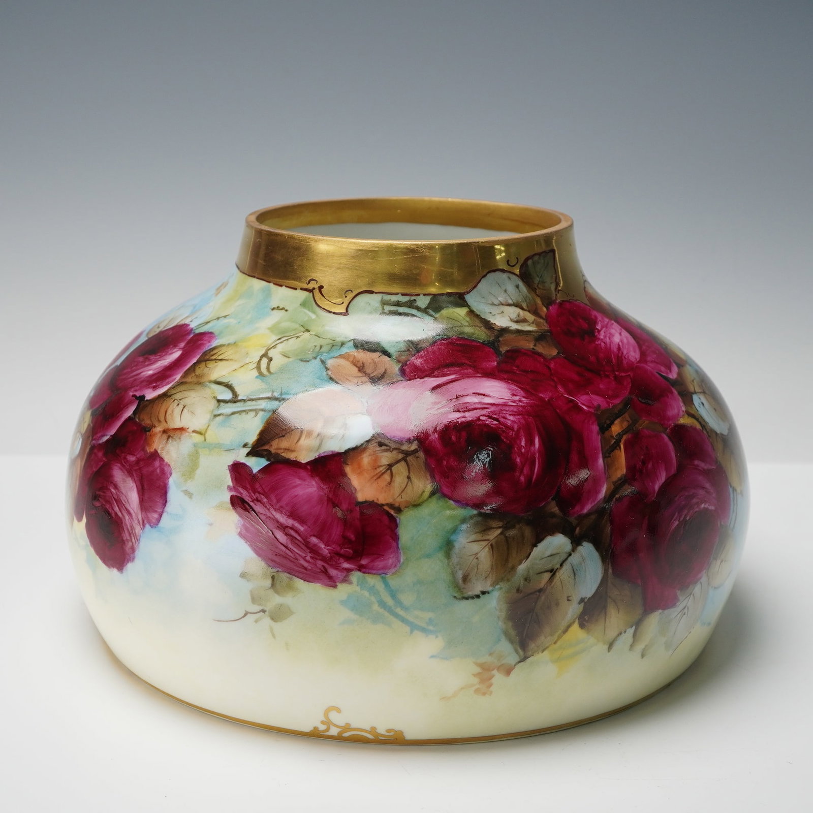M. Rost LeRoy Limoges Porcelain Floral Rose Vase: This Limoges porcelain vase has a rounded body with a wide flat bottom and a narrow opening finished with a gilded collar. It is hand painted with vivid red roses and soft foliage that wrap around the