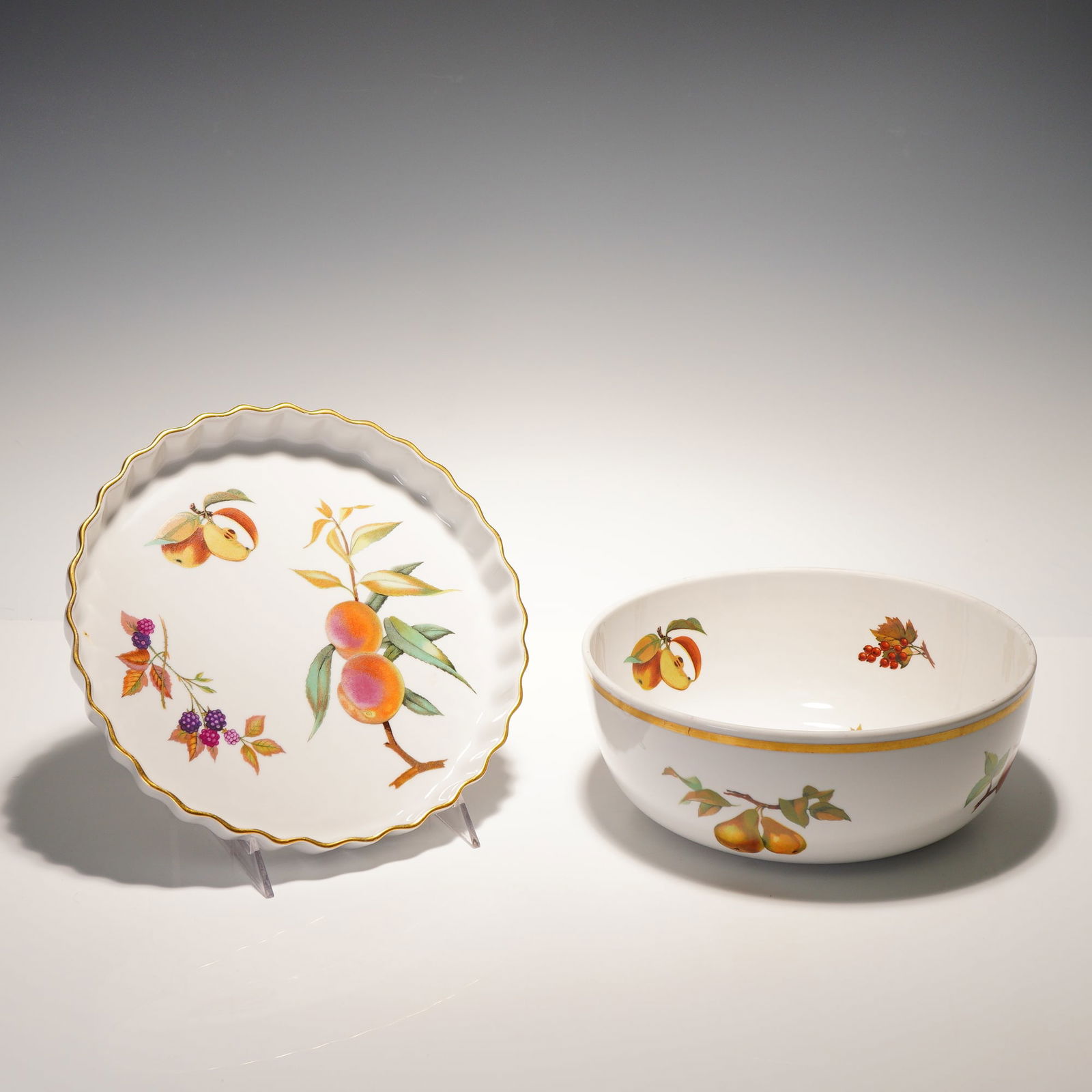 2pc Royal Worcester Porcelain Serveware, Evesham: A 2-piece porcelain serveware lot comprising one fluted quiche dish and one round salad serving bowl. Both feature the Evesham pattern with assorted fruit decoration and gold trim. Each piece is backs