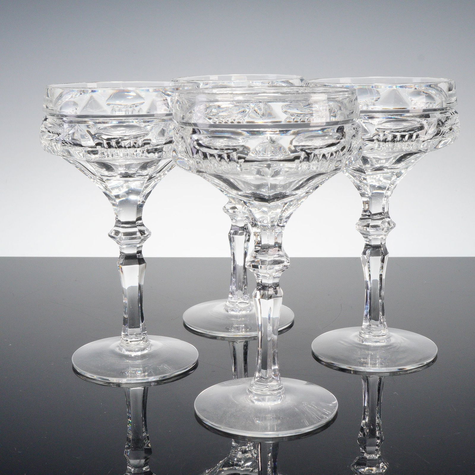 4pc Clear Cut Crystal Cocktail Coupe Glasses: Set of four clear cut crystal cocktail coupe glasses featuring faceted stems and geometric banding with fine prism detail. Elegant mid-century design, perfect for serving champagne or cocktails. Issue
