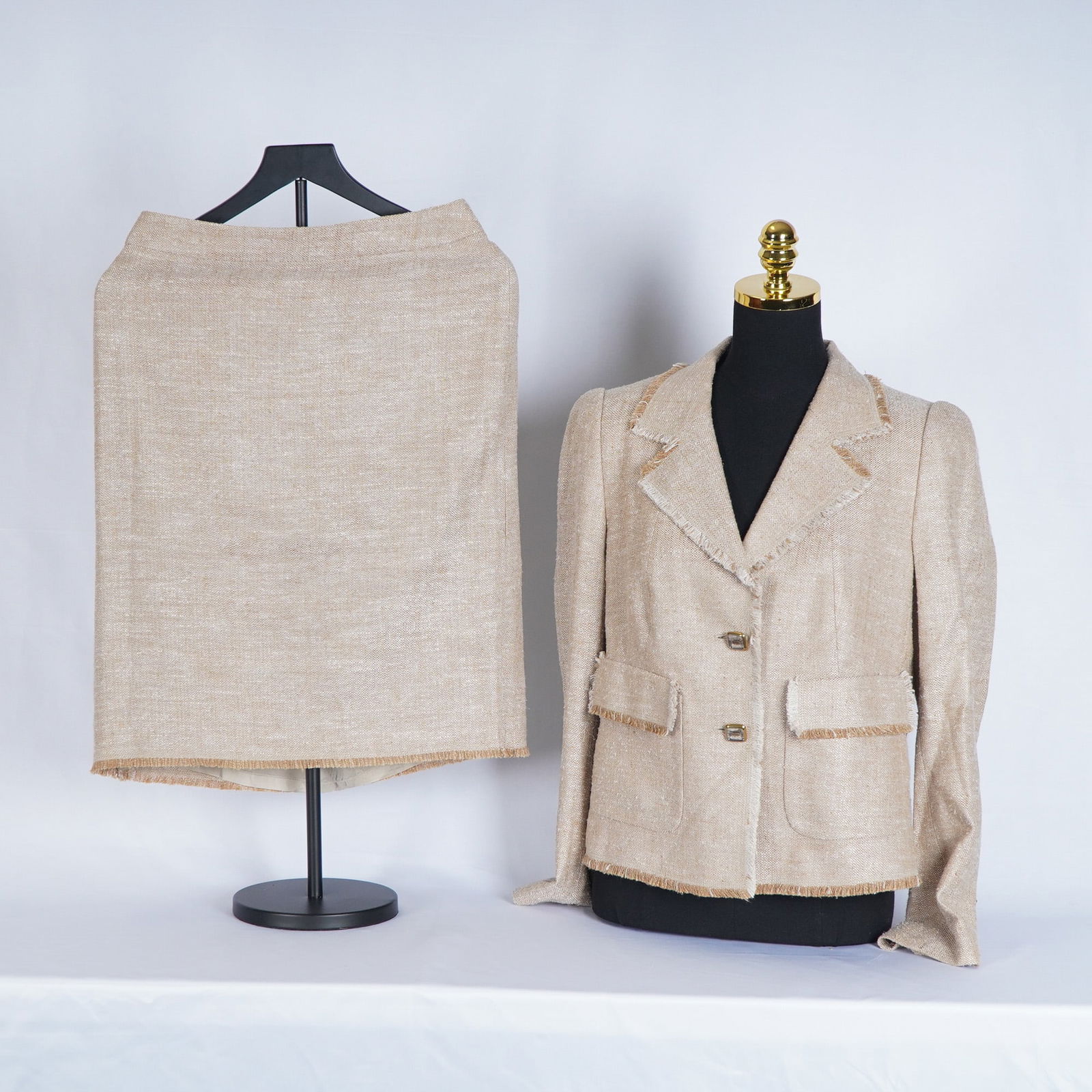 Escada Beige Tweed Skirt Suit: Tweed skirt suit featuring a tailored single-breasted jacket with notched lapels, two front flap pockets, button closures with the Escada name, and subtle fringe trim along the collar, hem, and pocket