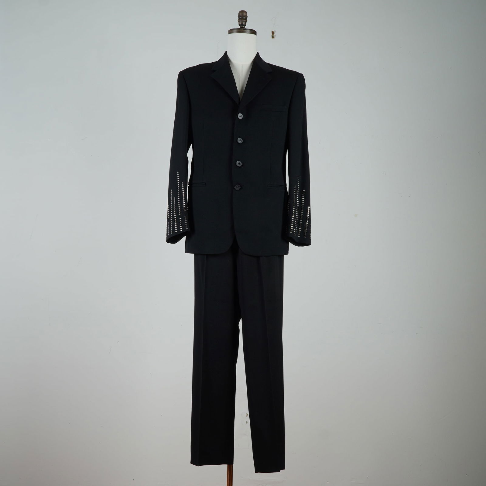 Gianni Versace Versus Black Studded Suit: A two piece pants and coat made of black wool. Made with notched lapels, structured shoulders, and decorative metallic stud embellishment arranged in descending linear patterns along the sleeves. The