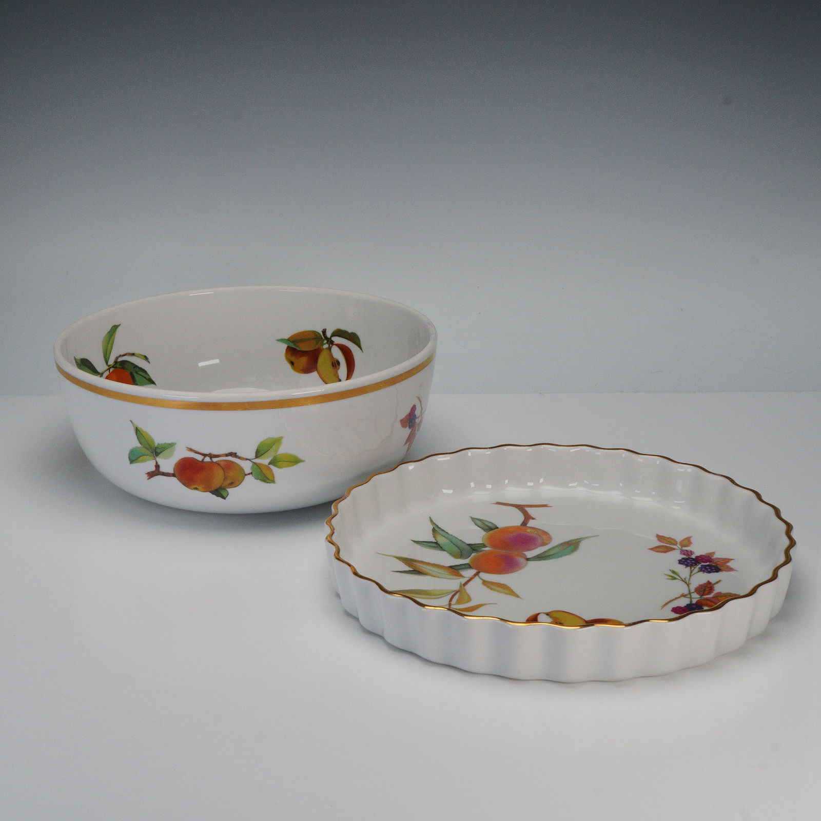 2pc Royal Worcester Porcelain Serveware, Evesham: A 2-piece porcelain serveware lot comprising one fluted quiche dish and one round salad serving bowl. Both feature the Evesham pattern with assorted fruit decoration and gold trim. Each piece is backs