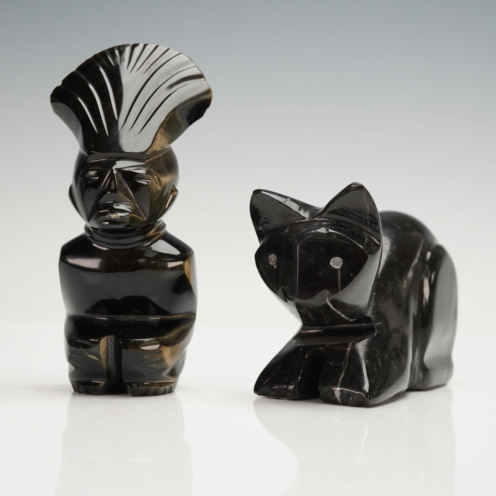 2pc Carved Obsidian Native American and Cat Figurines: A duo of carved obsidian figurines featuring a golden sheen standing Native American figure with a tall feathered headdress and a crouching cat with simple incised facial details. Dimensions: Largest