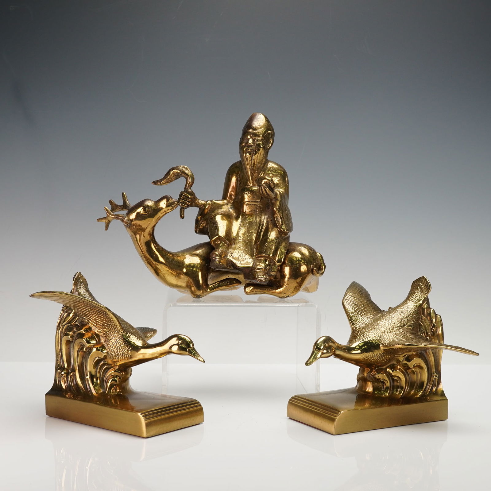 3pc Shou Xing and Flying Ducks Gilded Figurines and Bookends: This trio includes a gilded Shou Xing figure seated on a deer while holding a curved staff and wearing a detailed robe. It also includes two gilded duck bookends that show ducks in flight above styliz