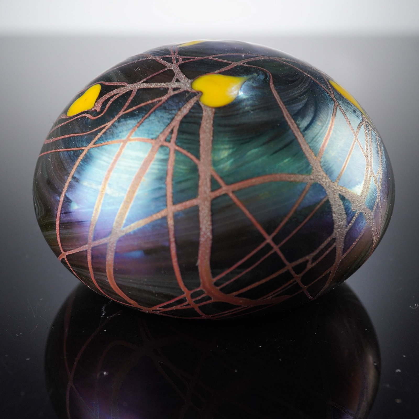 Orient & Flume Iridescent Art Glass Paperweight: An Orient & Flume iridescent art glass paperweight. The spherical form features a dark teal and purple iridescent ground displaying a subtle swirling pattern. The surface is adorned with an intricate