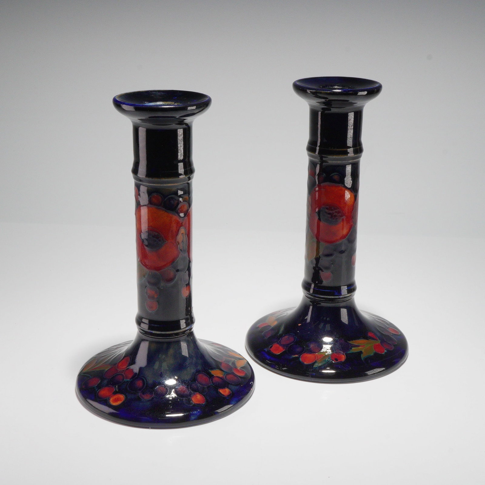 Pair of Moorcroft Pottery Candlesticks, Pomegranate: This pair of Moorcroft candlesticks presents the tube-lined Pomegranate pattern with cobalt grounds and hand-painted fruit and foliage. The decoration shows the characteristic segmented pomegranate fo