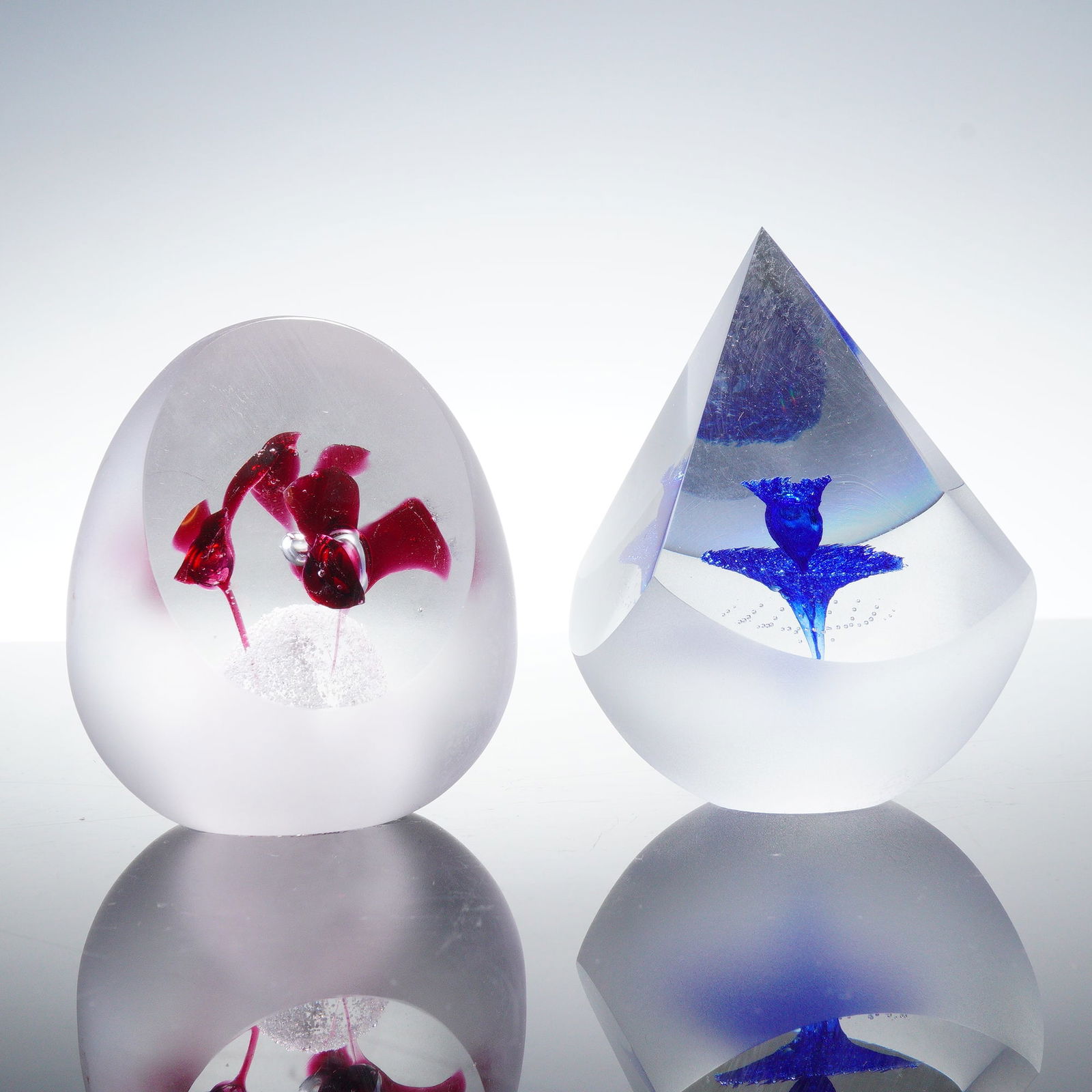 2pc Art Glass Paperweights, Red and Blue Floral Designs: A group of two art glass paperweights, one featuring a red flower encased within a rounded clear form with frosted exterior, the other with a blue flower enclosed in a teardrop-shaped clear body with