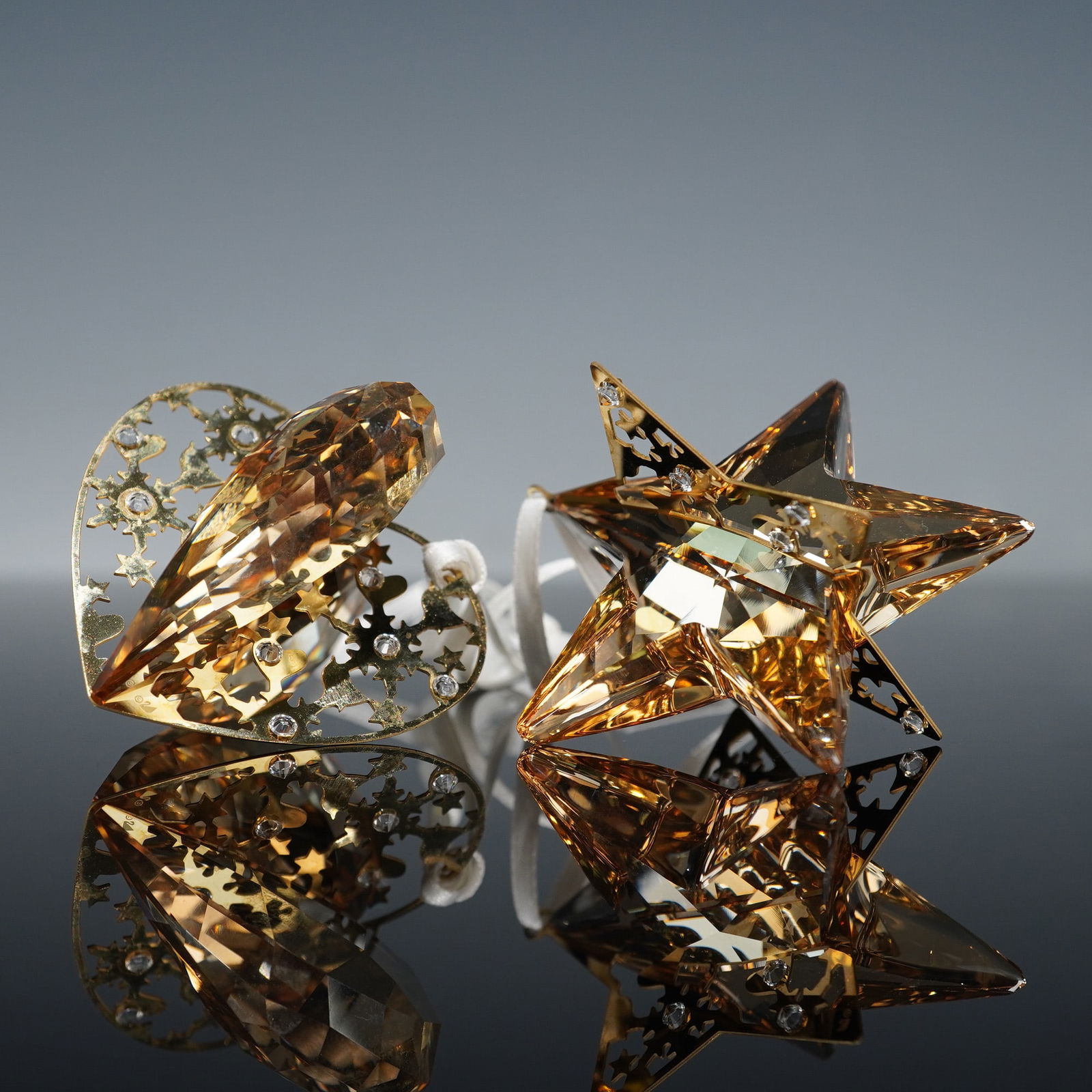 Pair of Swarovski Golden Shadow Crystal and Metal Ornaments: This is a 2pc Swarovski crystal and metal ornaments set, featuring one heart-shaped and one five-pointed star-shaped ornament. Both pieces are constructed from faceted, amber-toned crystal (identified