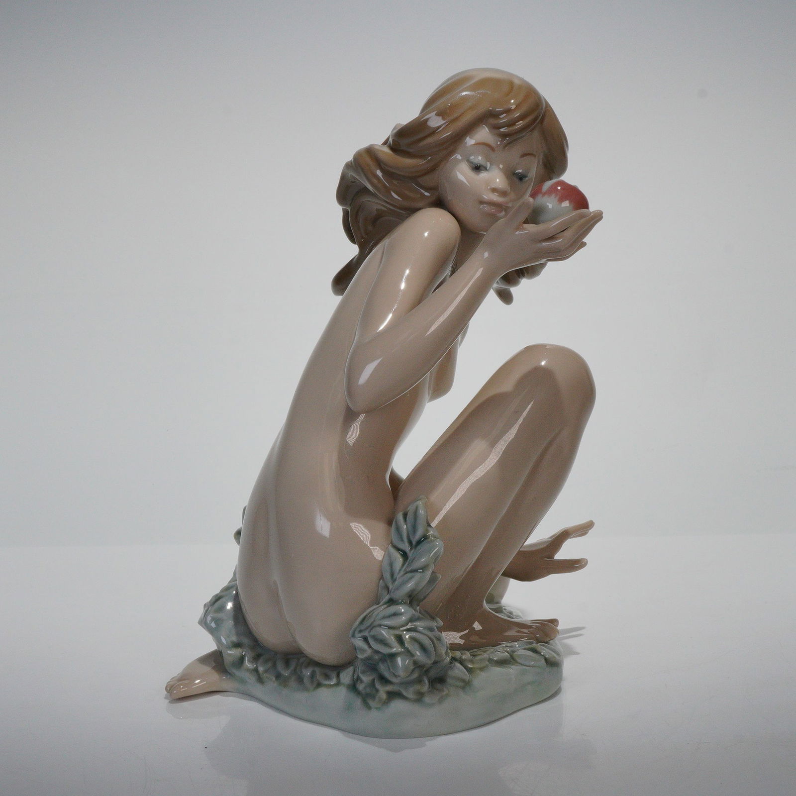 Lladro Porcelain Figurine, Eve 1001482: Depicts a nude female figure in a crouching pose, holding an apple in her right hand and gazing downwards. The figure has brown hair, and the naturalistic base features relief-molded foliage in a pale