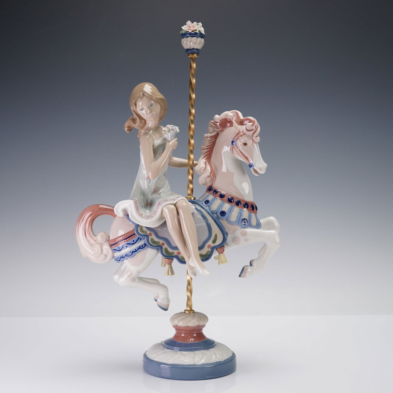 Lladro Porcelain Sculpture, Girl on Carousel 01001469: Sculpture depicting a young girl seated gracefully atop an ornate carousel horse. Designed in Lladro's classic glossy finish, the piece showcases pastel blue, pink, and ivory tones with intricate sadd