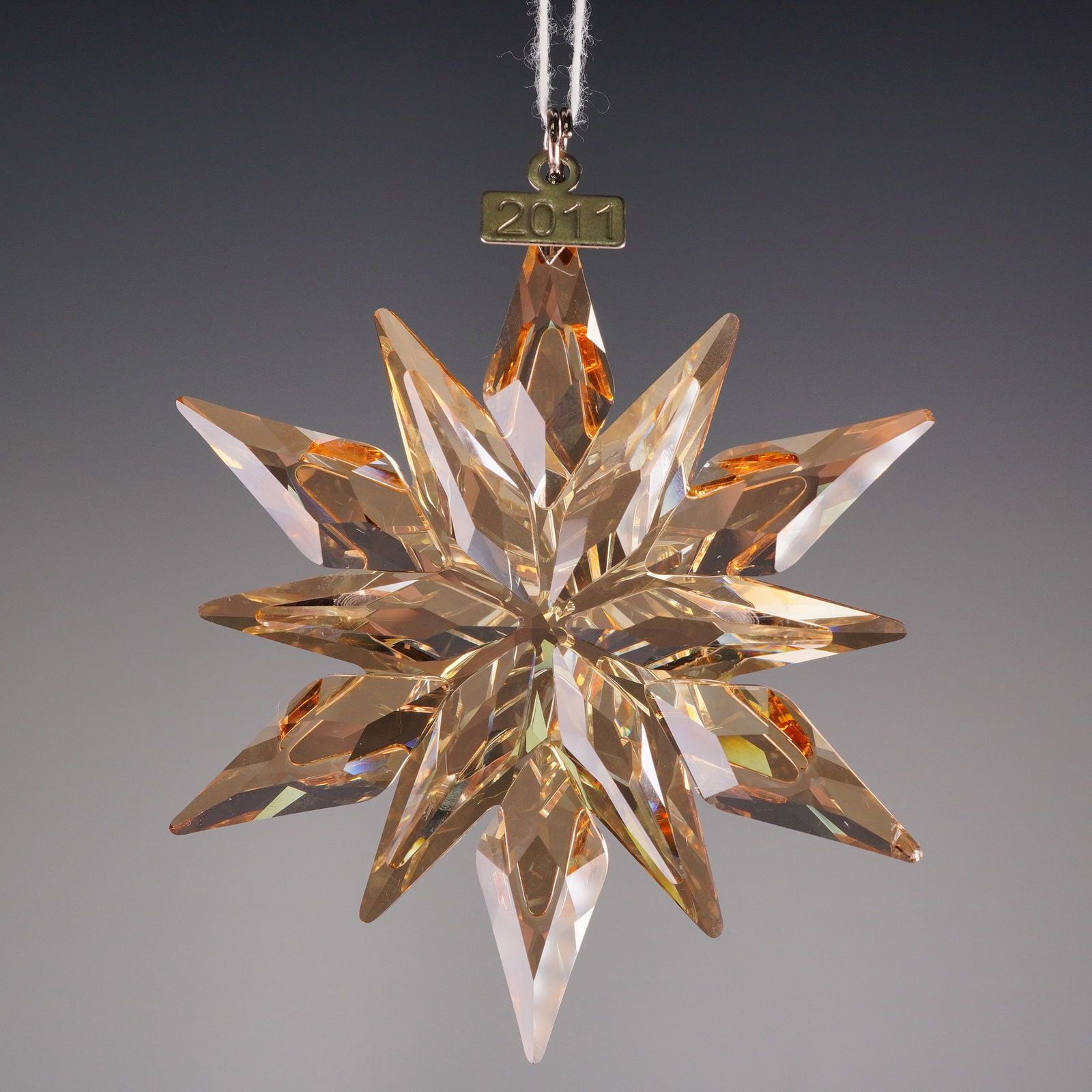 Swarovski SCS 2011 Annual Edition Gold Christmas Star Ornament: A Swarovski SCS 2011 Annual Edition Gold Christmas Ornament (model 1092040). The piece is fashioned from highly faceted cut crystal, exhibiting 12 pointed snowflake with a gold hue. The silver toned t