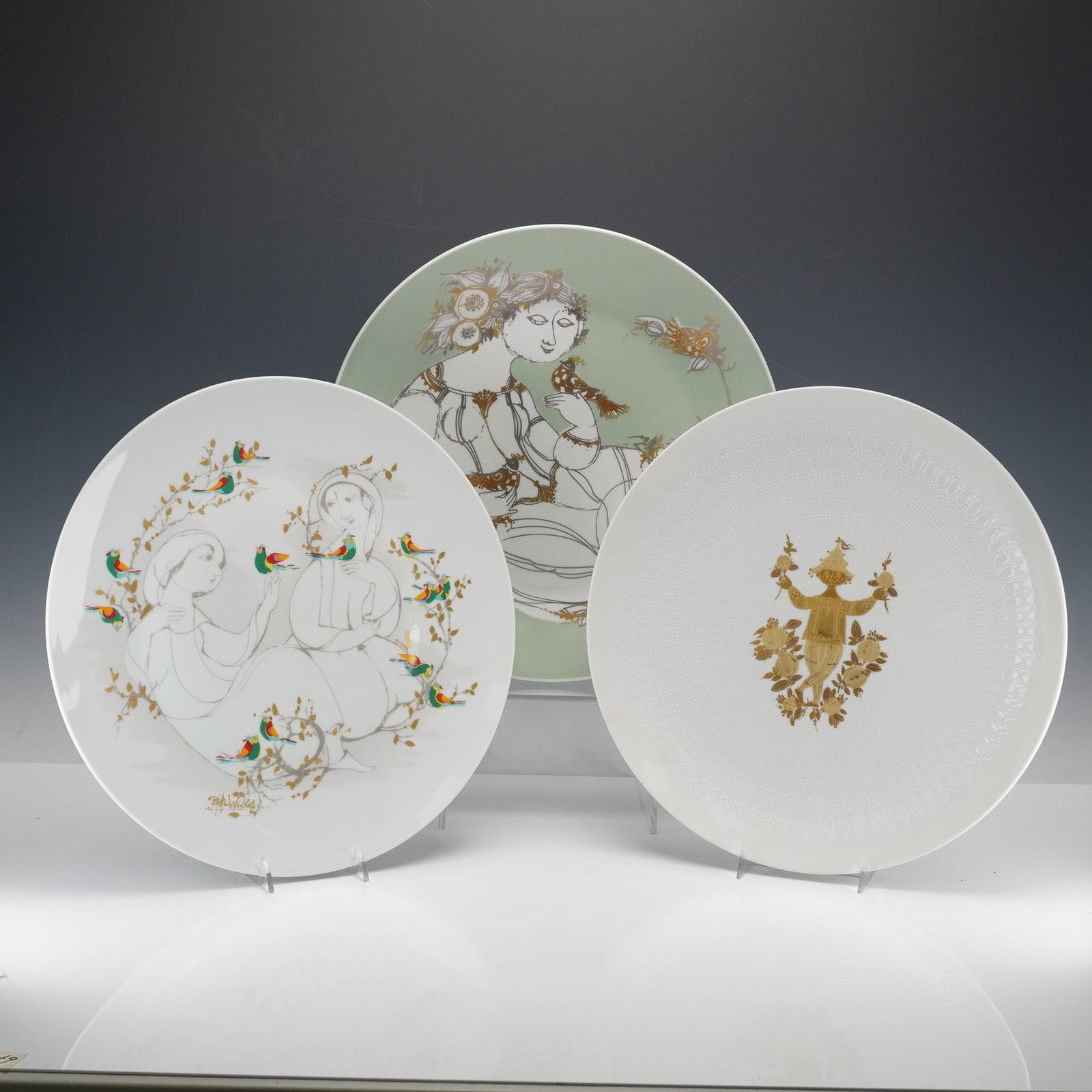 3pc Rosenthal Studio Linie by Bjorn Wiinblad Porcelain Decorative Plates: The plates depict stylized figures of women with birds and foliage; two on a white ground with gold and polychrome accents and another on a pale green ground with gold and grey accents. Two plates are